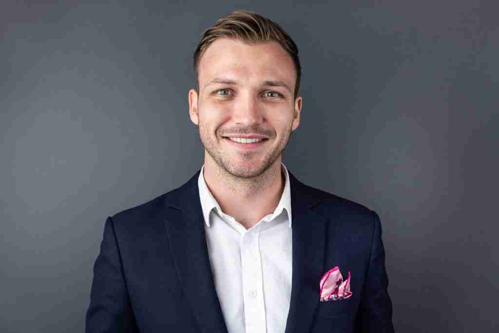 A man in a dark suit and white shirt is smiling at the camera. He has short hair and a pink pocket square. The background is a plain, dark gray color.