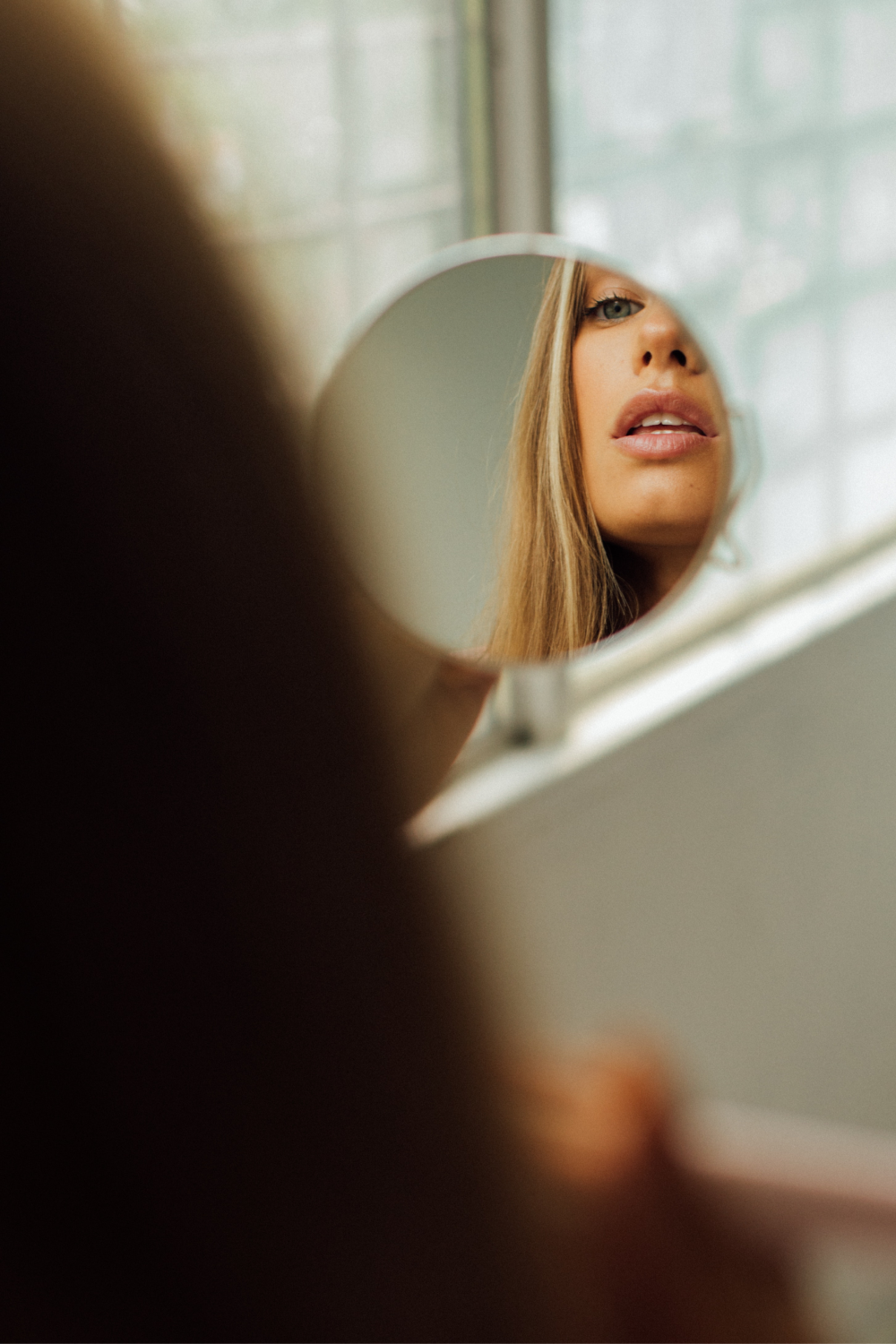 A woman holds a small round mirror, reflecting her lips and part of her face. She has long hair, and the background shows a blurred window with a soft, natural light.