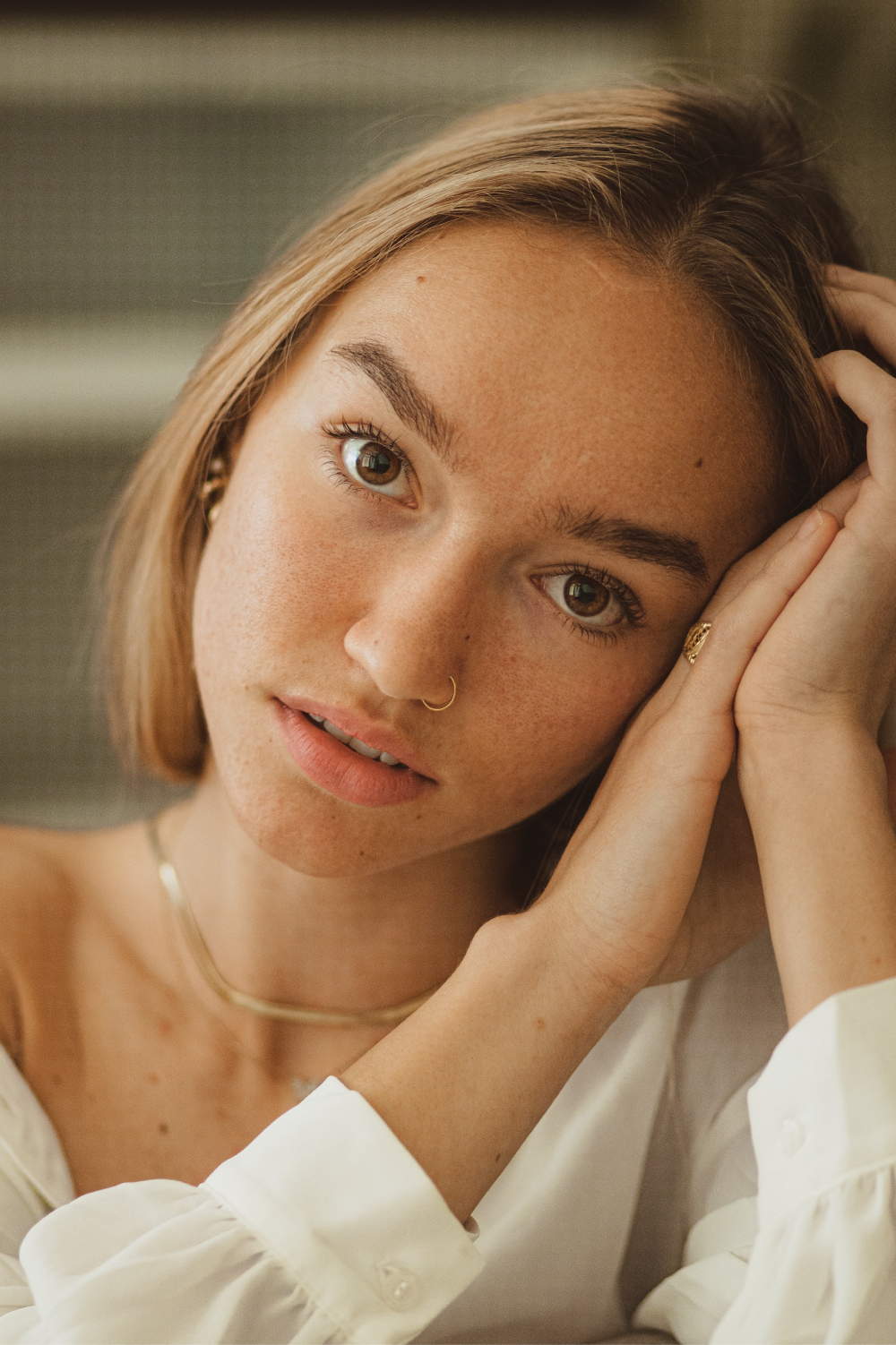 A person with light brown hair and wearing a white shirt leans their head on their hands. They have a nose ring, earrings, and a necklace. The expression is relaxed and the background is blurred.