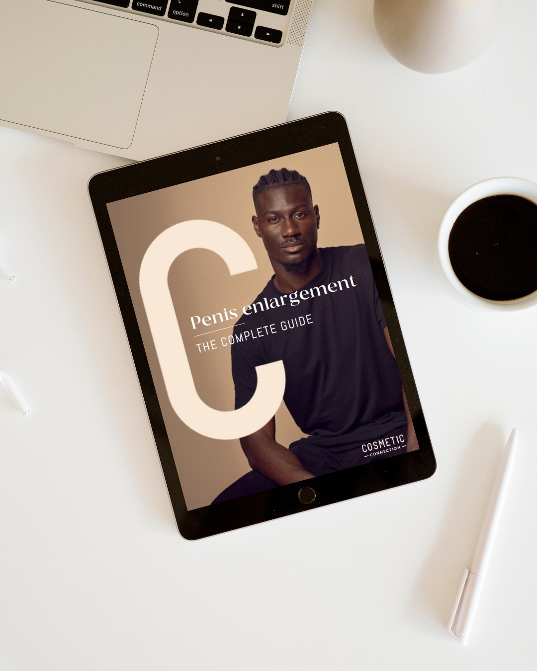 A tablet on a white desk displays an e-book titled Penis enlargement: The Complete Guide with a photo of a man. Nearby are part of a laptop, a cup of coffee, a pen, and a round object.