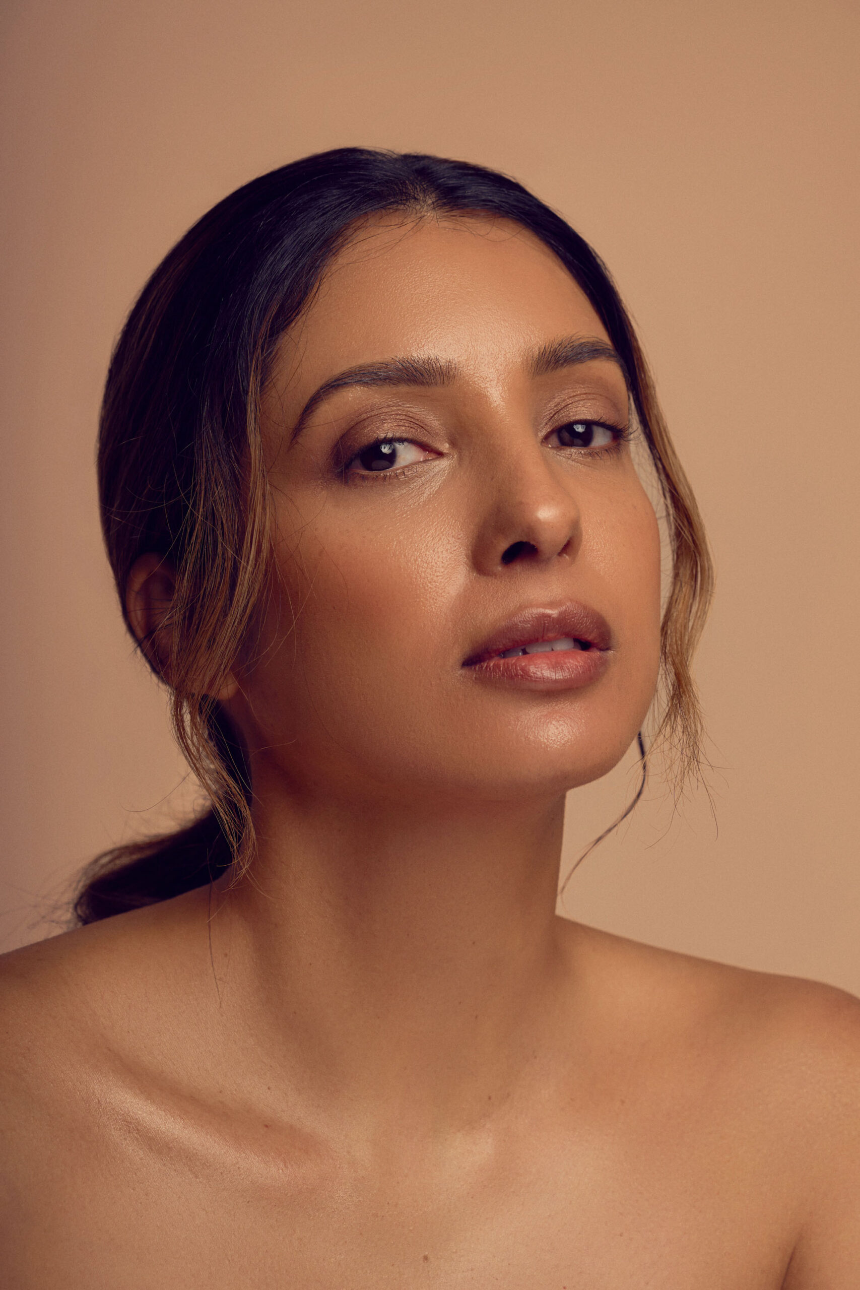 A woman with long dark hair pulled back, showcasing a natural makeup look. Her shoulders are bare, and she is looking directly at the camera against a neutral background.