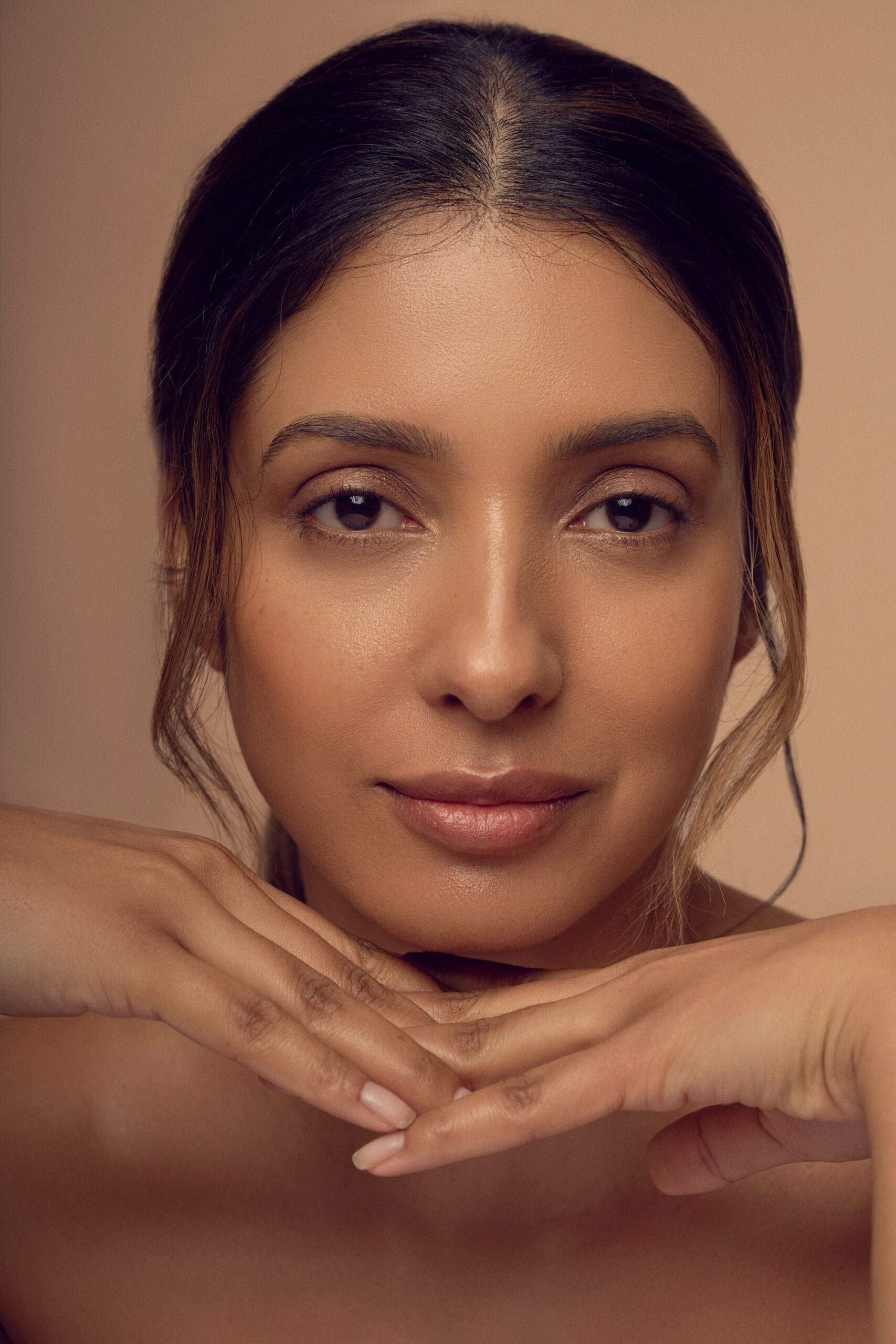 A woman with long brown hair and subtle makeup poses against a beige background. She gazes directly at the camera with a serene expression, resting her chin on her hands with fingers crossed.