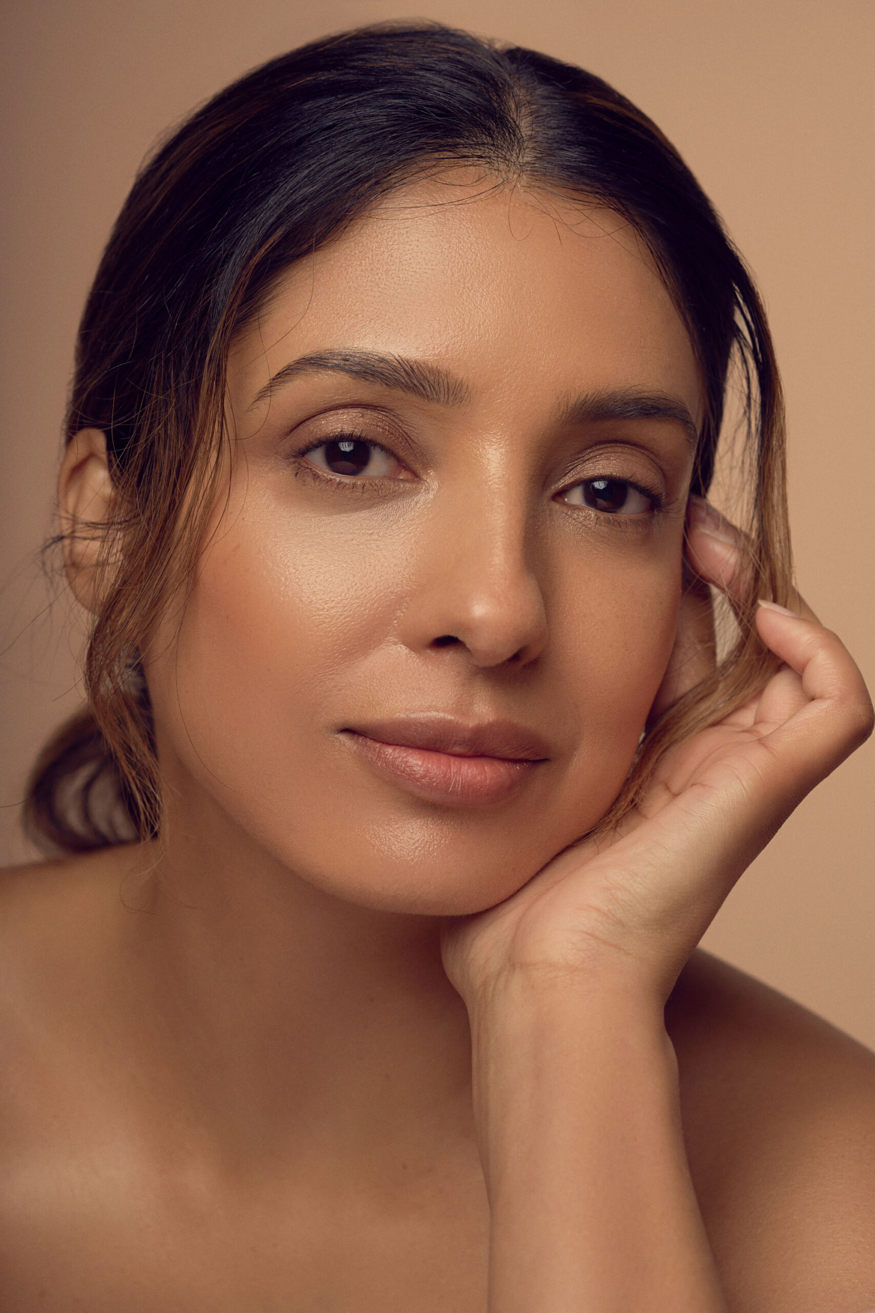 A woman with shoulder-length hair gazes softly at the camera, resting her chin on her hand. She wears natural makeup and is positioned against a plain, neutral background.