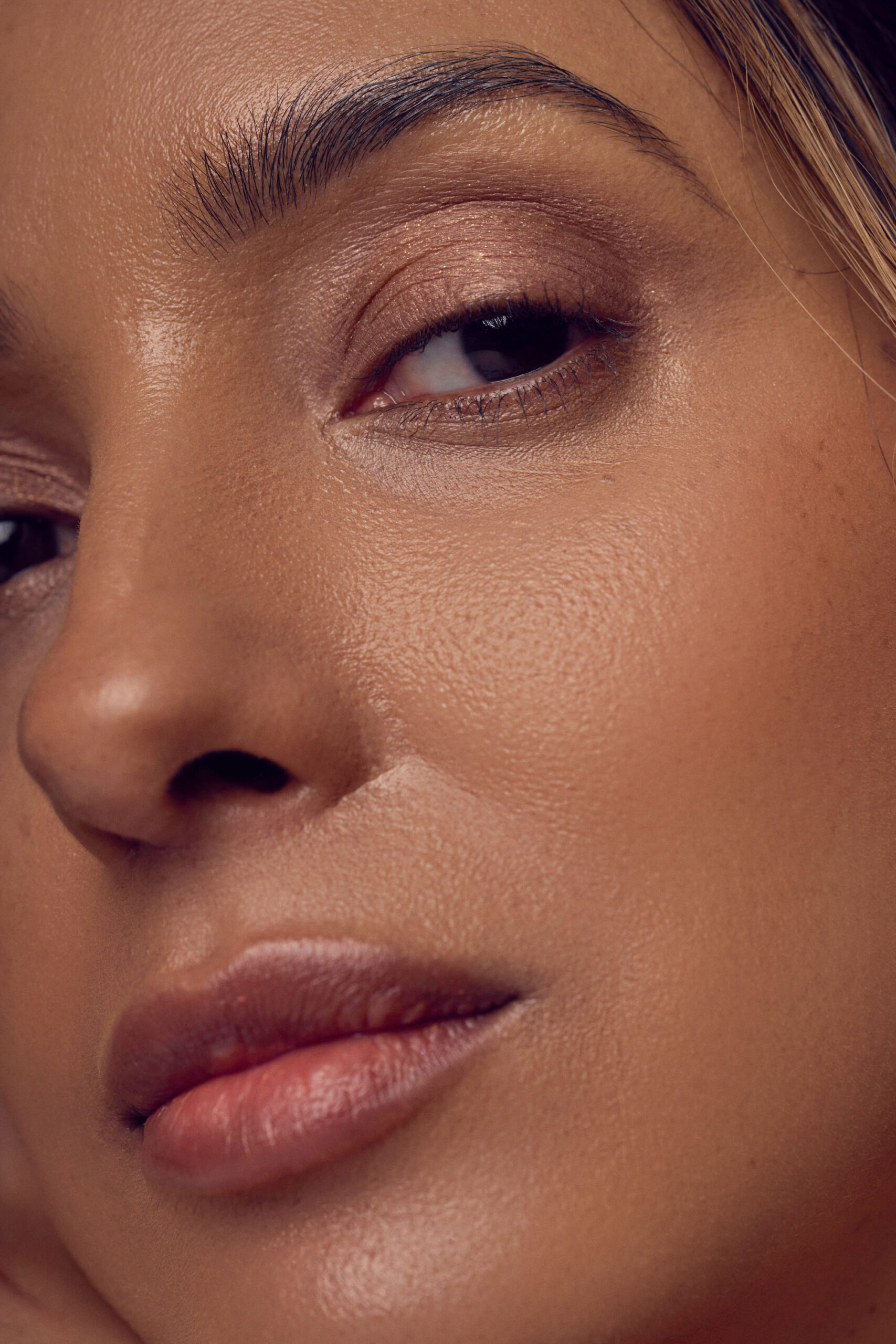 Close-up of a person with smooth, glowing skin. Their eye is softly closed, and they have natural makeup with neutral tones on their lips and eyelid. The image highlights the texture and radiance of their skin.