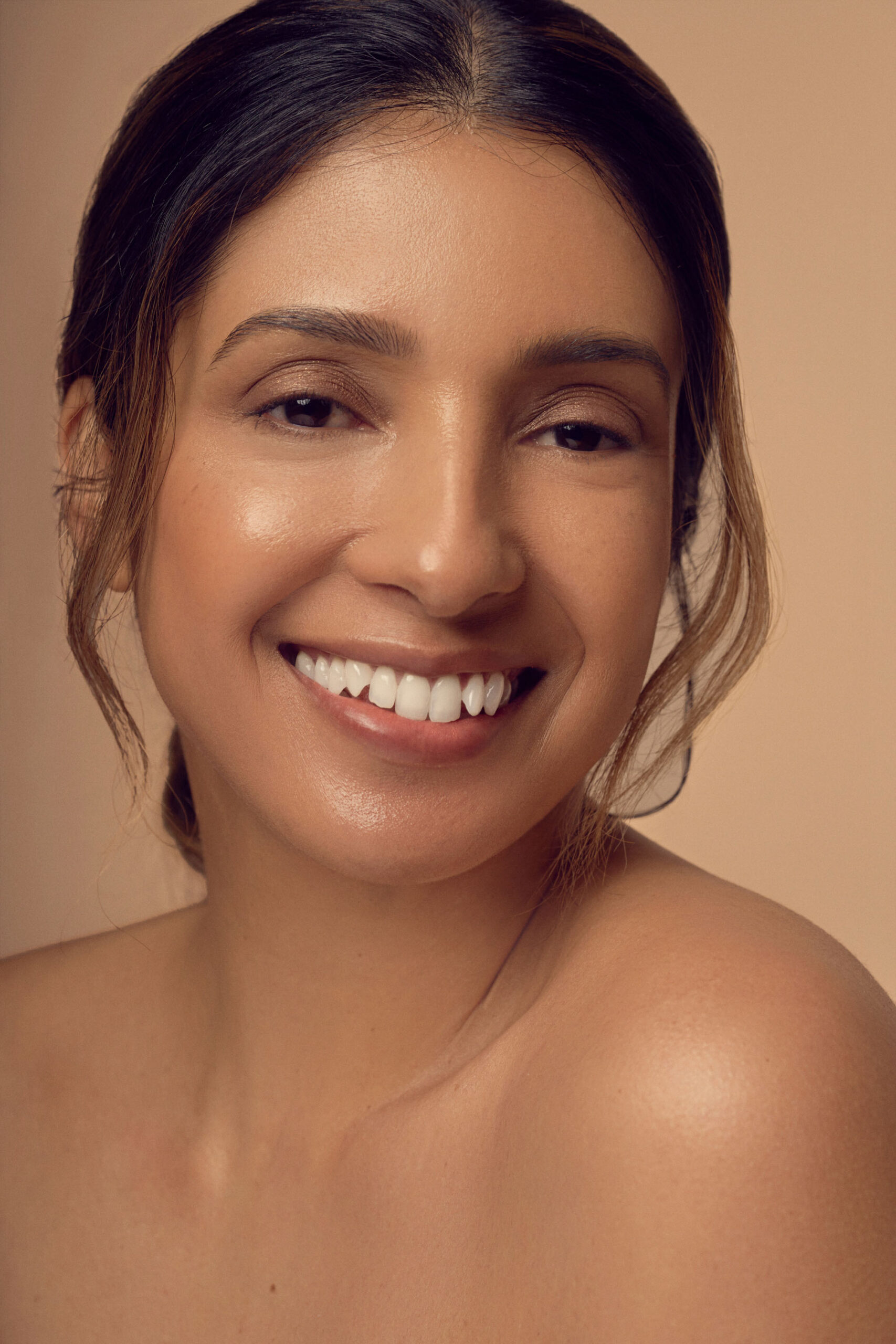 A person with long dark hair smiles warmly against a neutral background. They have glowing skin and visible dimples, creating an inviting and pleasant expression. Their shoulders are bare, adding to the overall natural and serene look.