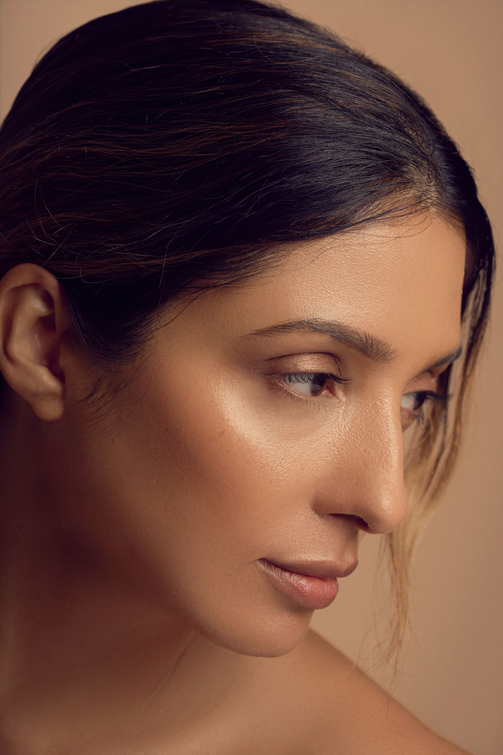 Close-up of a person with long hair tied back, showcasing flawless skin and soft makeup. Their head is turned slightly to the side, highlighting their profile against a neutral background.