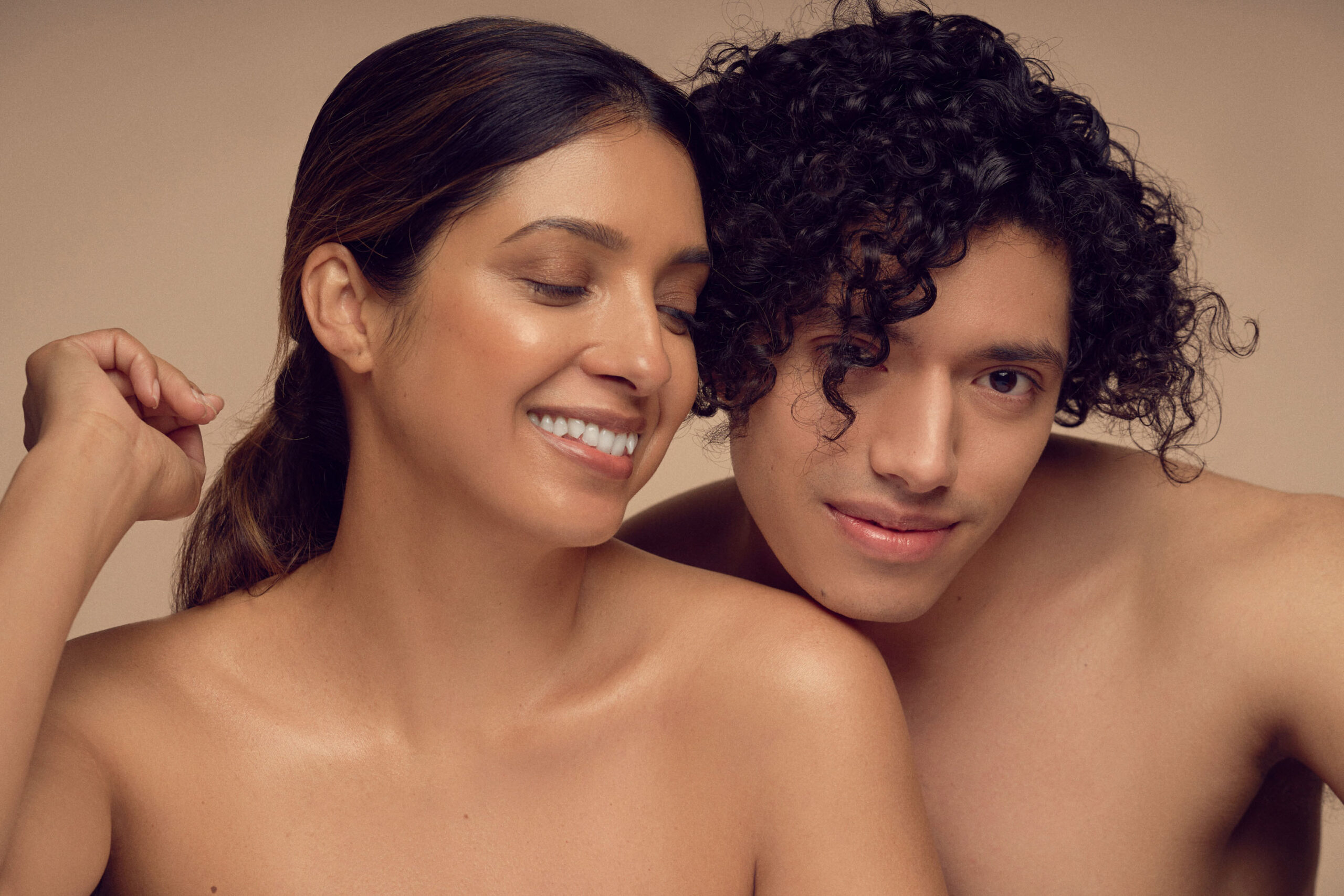 A close-up of two smiling individuals with bare shoulders against a neutral background. The person on the left has long dark hair pulled back, while the person on the right has short curly hair. Both have a relaxed and content expression.