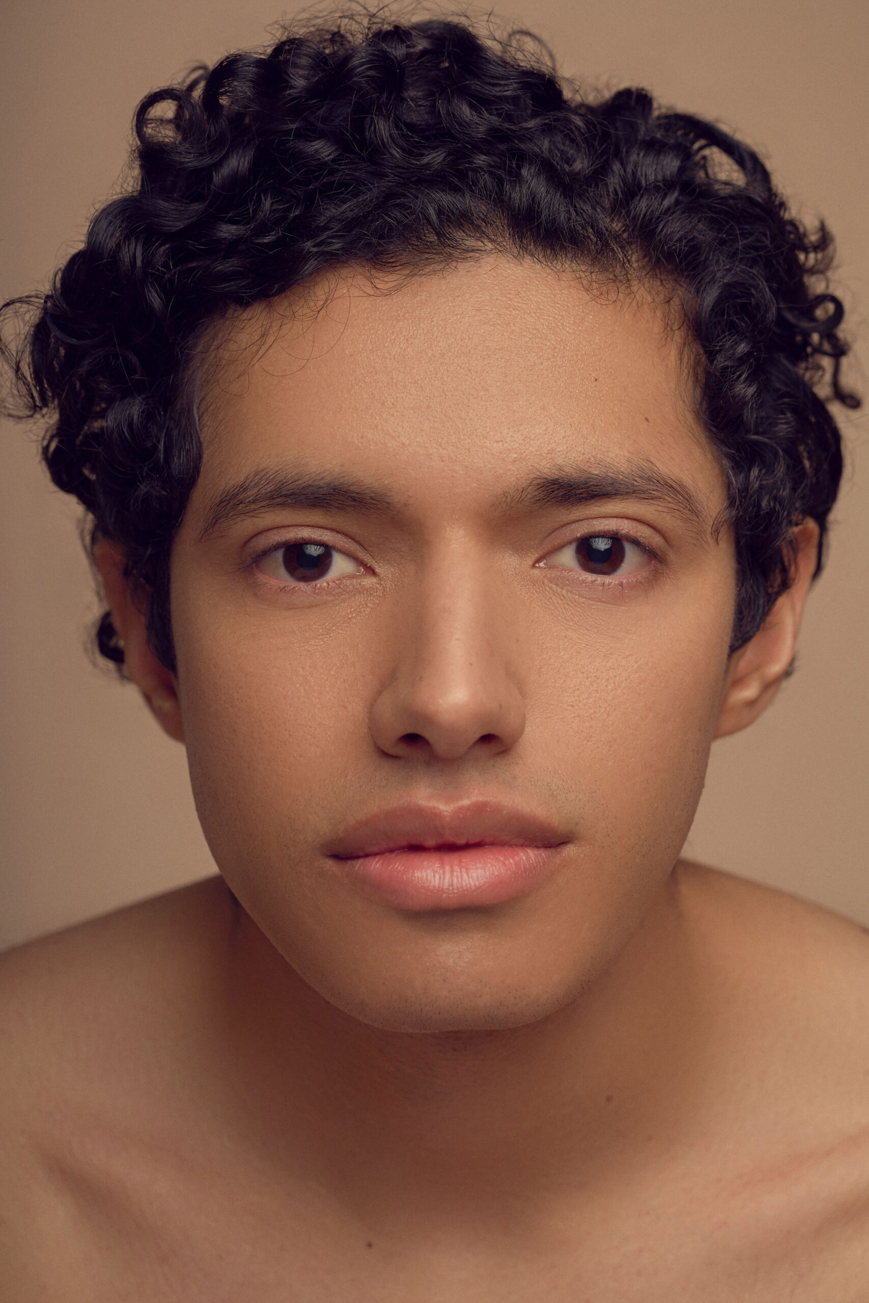 A person with short curly hair and clear skin looks directly into the camera with a neutral expression. The background is a soft beige color, complementing the soft lighting on the subjects face.