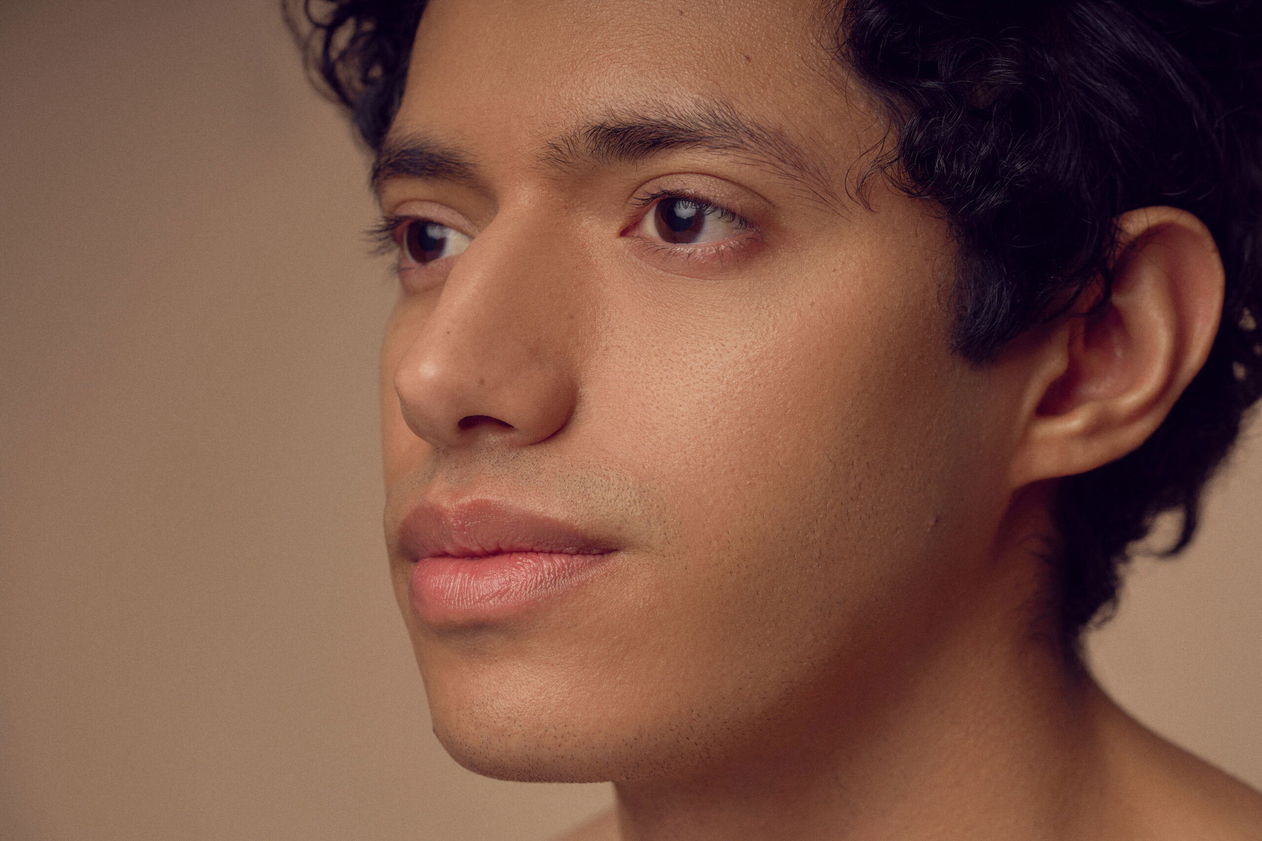 Close-up of a person with short, curly dark hair and smooth skin, gazing thoughtfully to the side against a soft, neutral background.