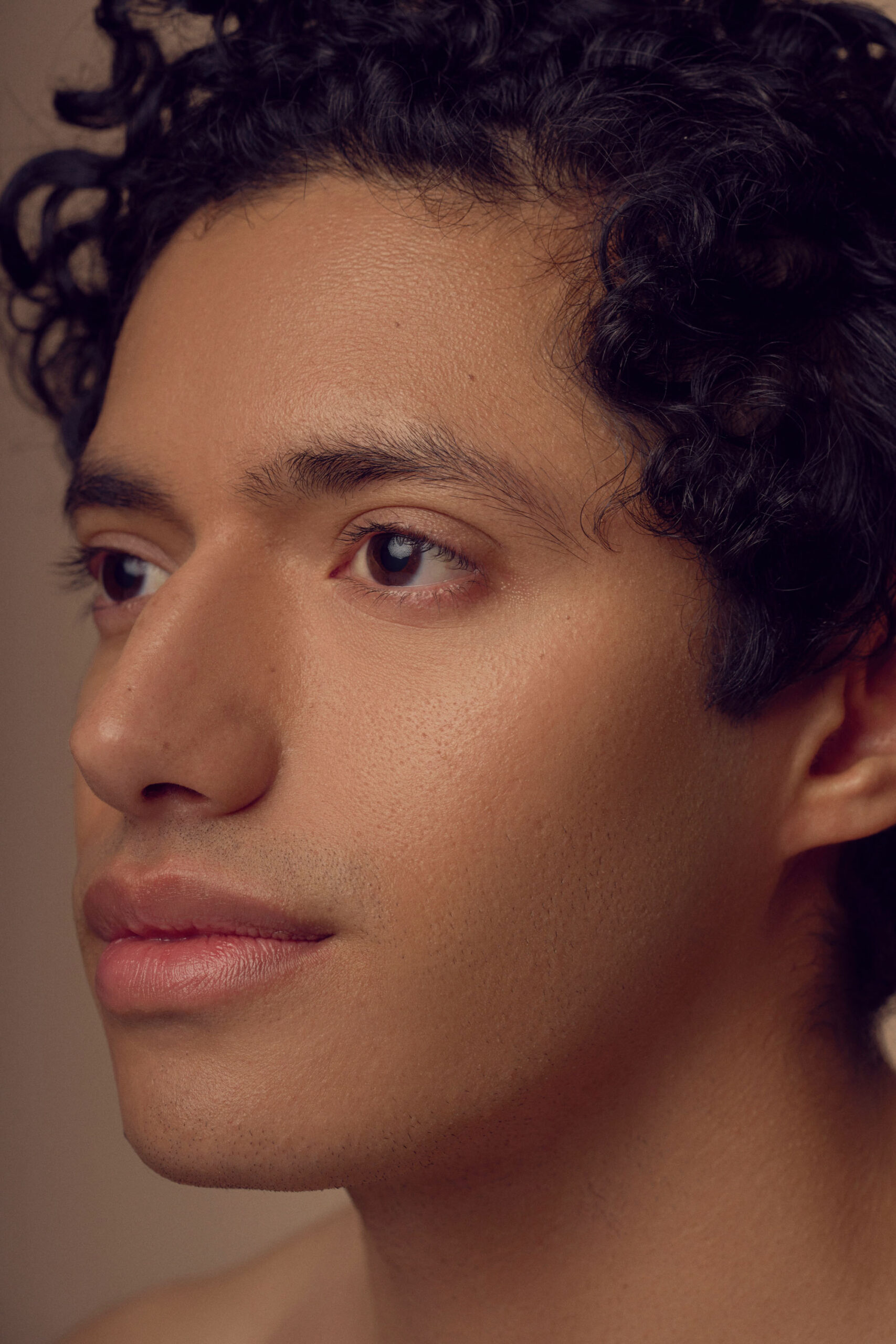 A person with curly hair and smooth skin gazes thoughtfully to the side. The close-up image highlights their serene expression and soft lighting.