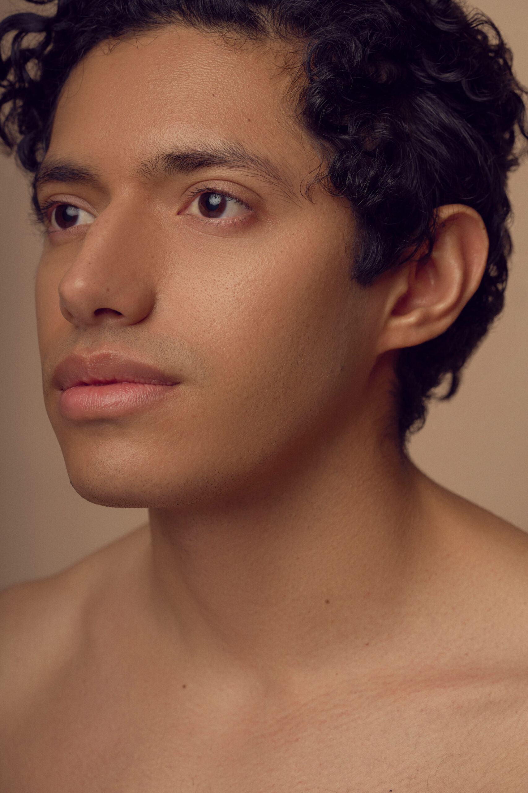 A close-up portrait of a person with curly dark hair. Their expression is calm, and they are looking into the distance against a neutral background. They have smooth skin and are not wearing a shirt.