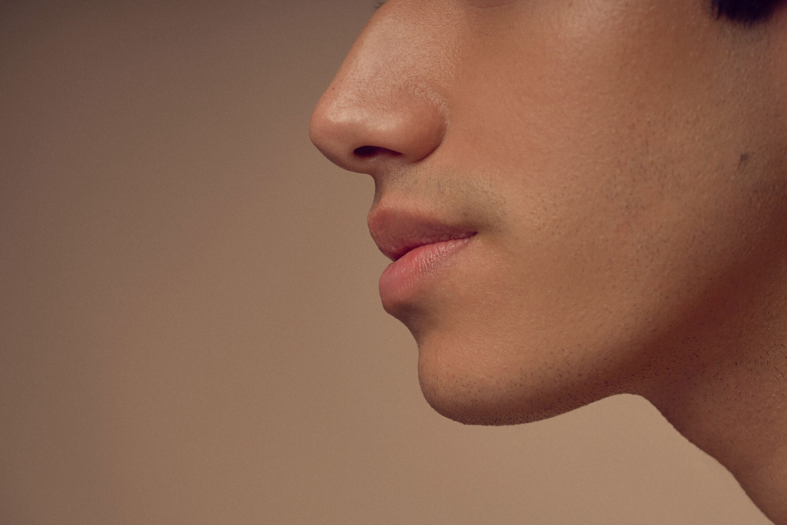 Close-up of a persons profile showing the lower half of their face, including their nose, lips, and chin. The background is blurred and neutral in color, highlighting the smooth complexion and subtle features.