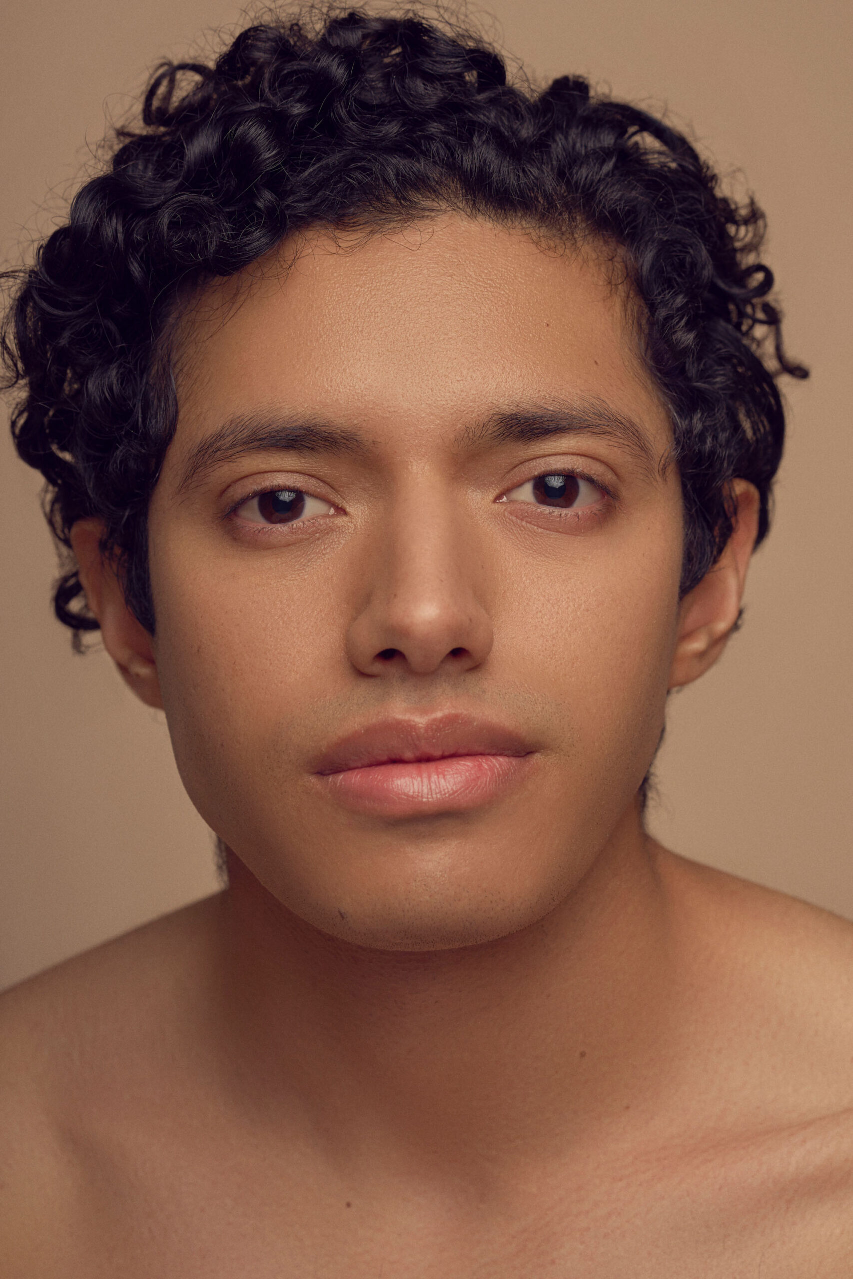 A person with short, curly hair and a neutral expression looks directly at the camera against a plain background. The image captures their face and shoulders.