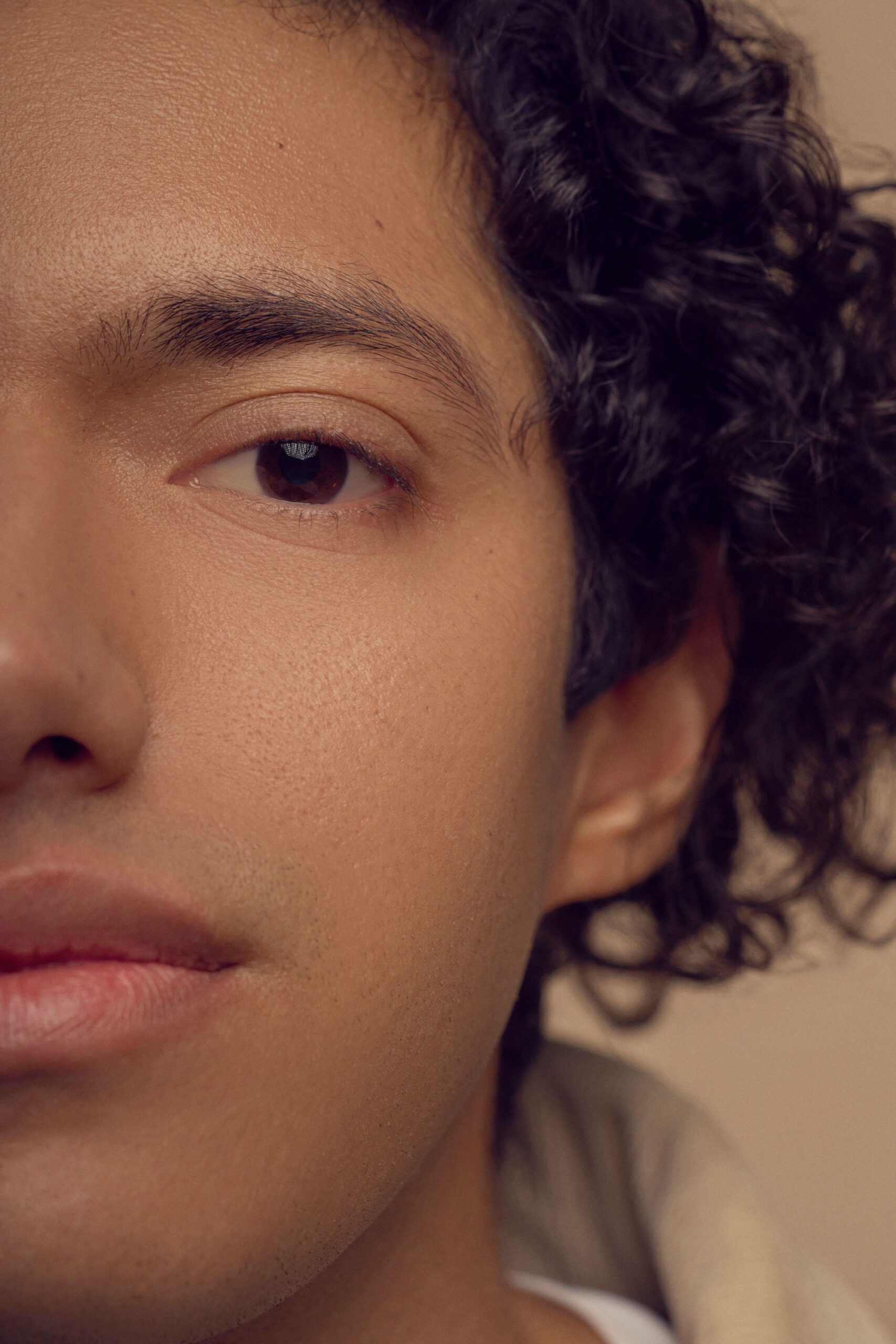 Close-up image of a person with curly hair and a neutral expression. Only the left side of their face is visible, highlighting one eye, half of the mouth, part of the cheek, and ear. The background is a soft beige color.