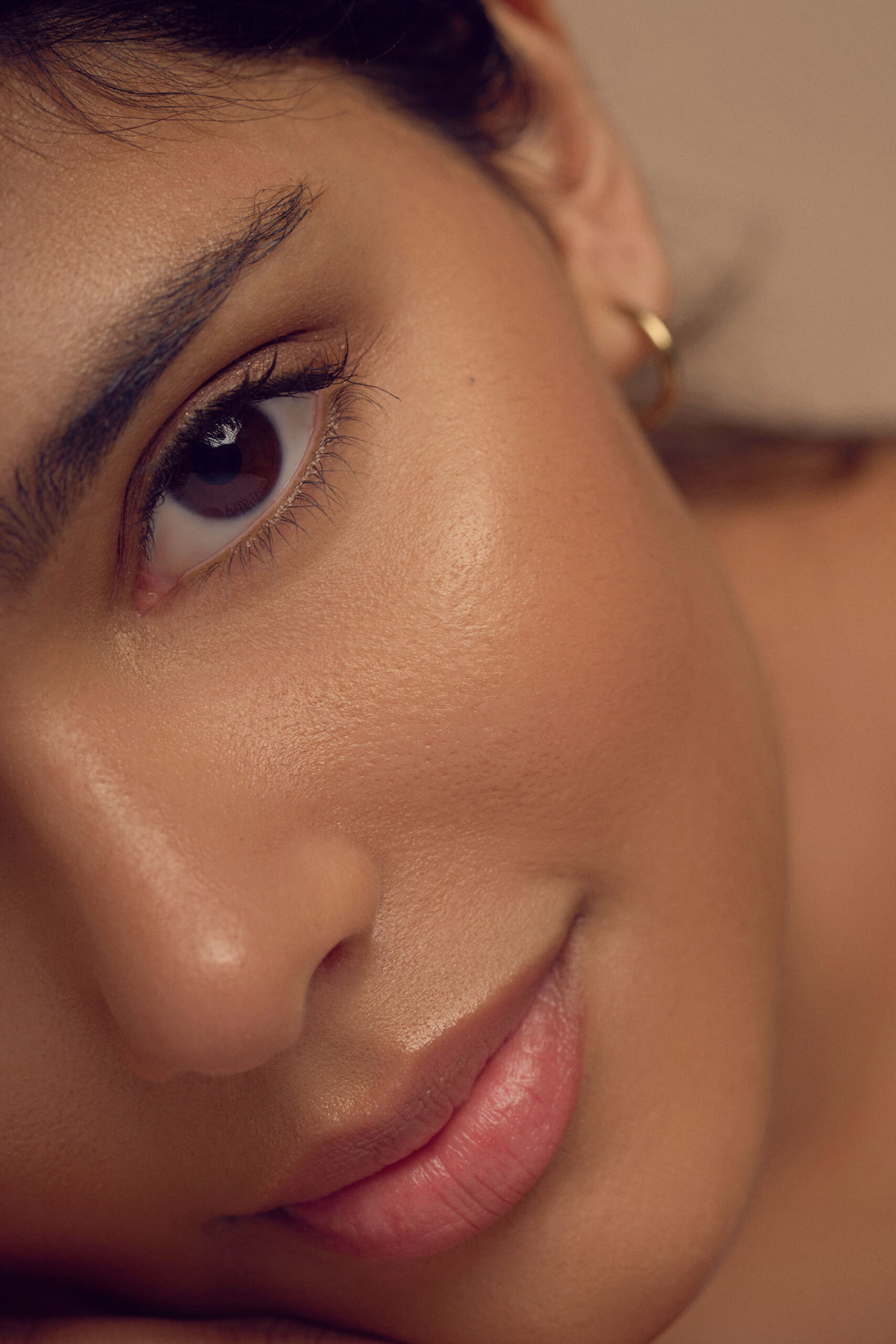 Close-up of a persons face showing smooth skin, a brown eye, and soft pink lips. The lighting is warm, highlighting the texture and natural beauty of the skin. The person wears a small hoop earring.