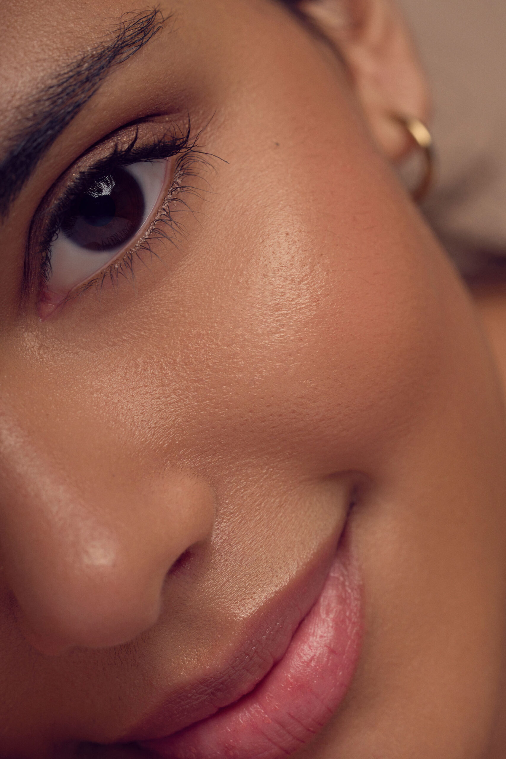 Close-up of a persons face focusing on their eye and cheek. The skin appears smooth and radiant, with natural makeup. They are smiling slightly, and a small gold hoop earring is visible.