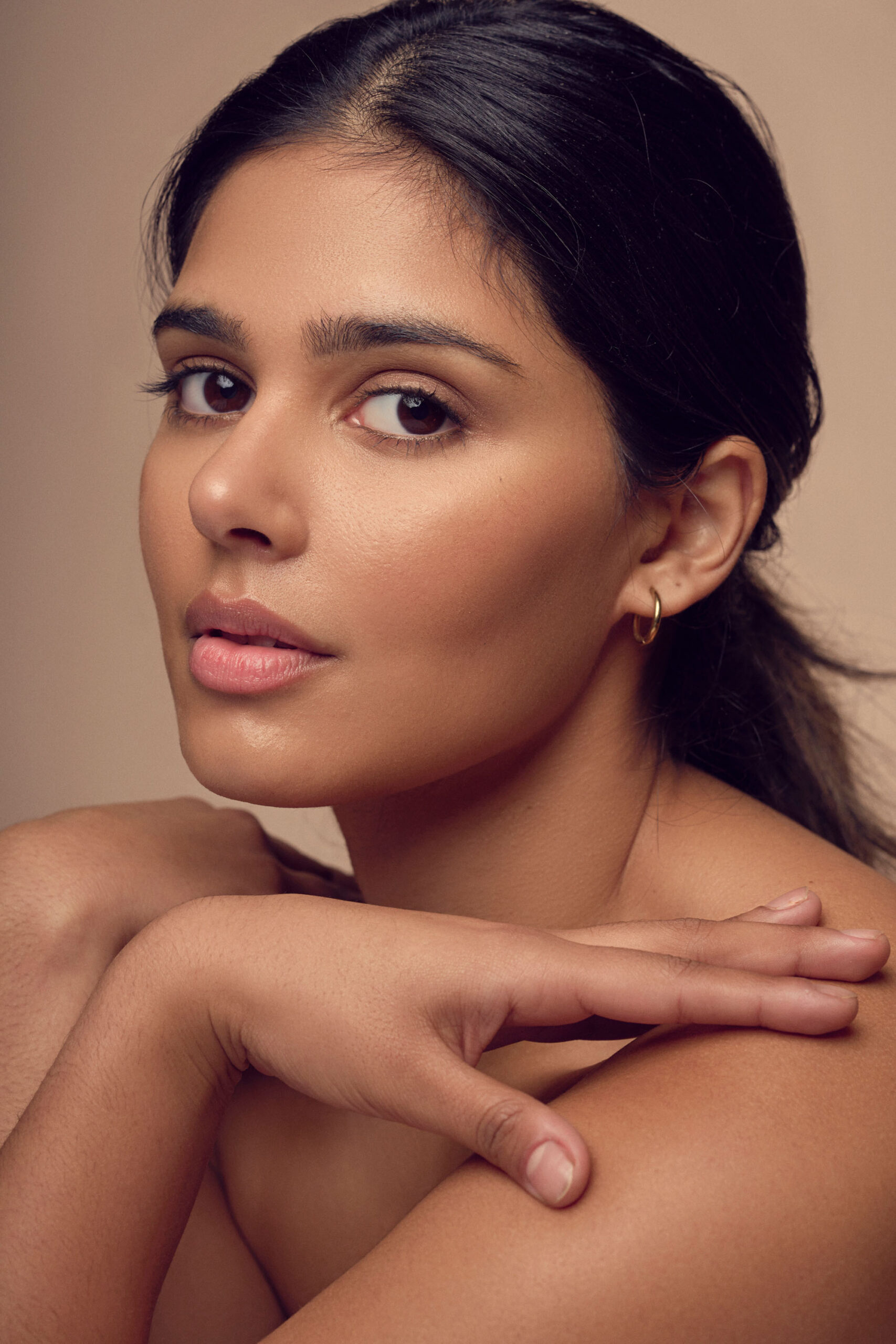 A woman with dark hair pulled back gazes at the camera. Her skin is smooth and she wears subtle makeup. Her hands are delicately crossed in front of her shoulders. The background is a soft neutral tone.