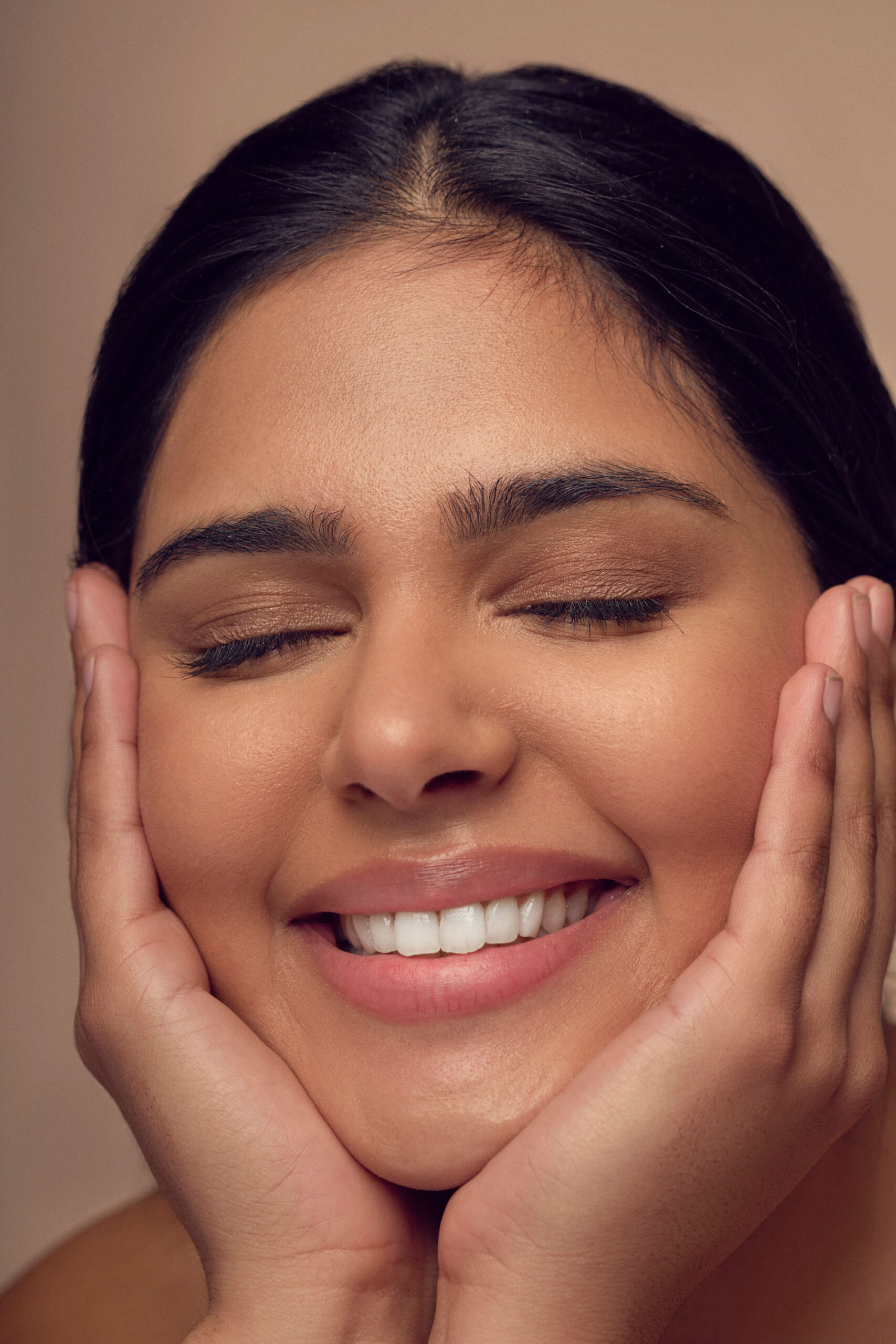 A person with dark hair smiling with eyes closed, gently holding their face with both hands. The background is a soft beige color, creating a warm and serene atmosphere.