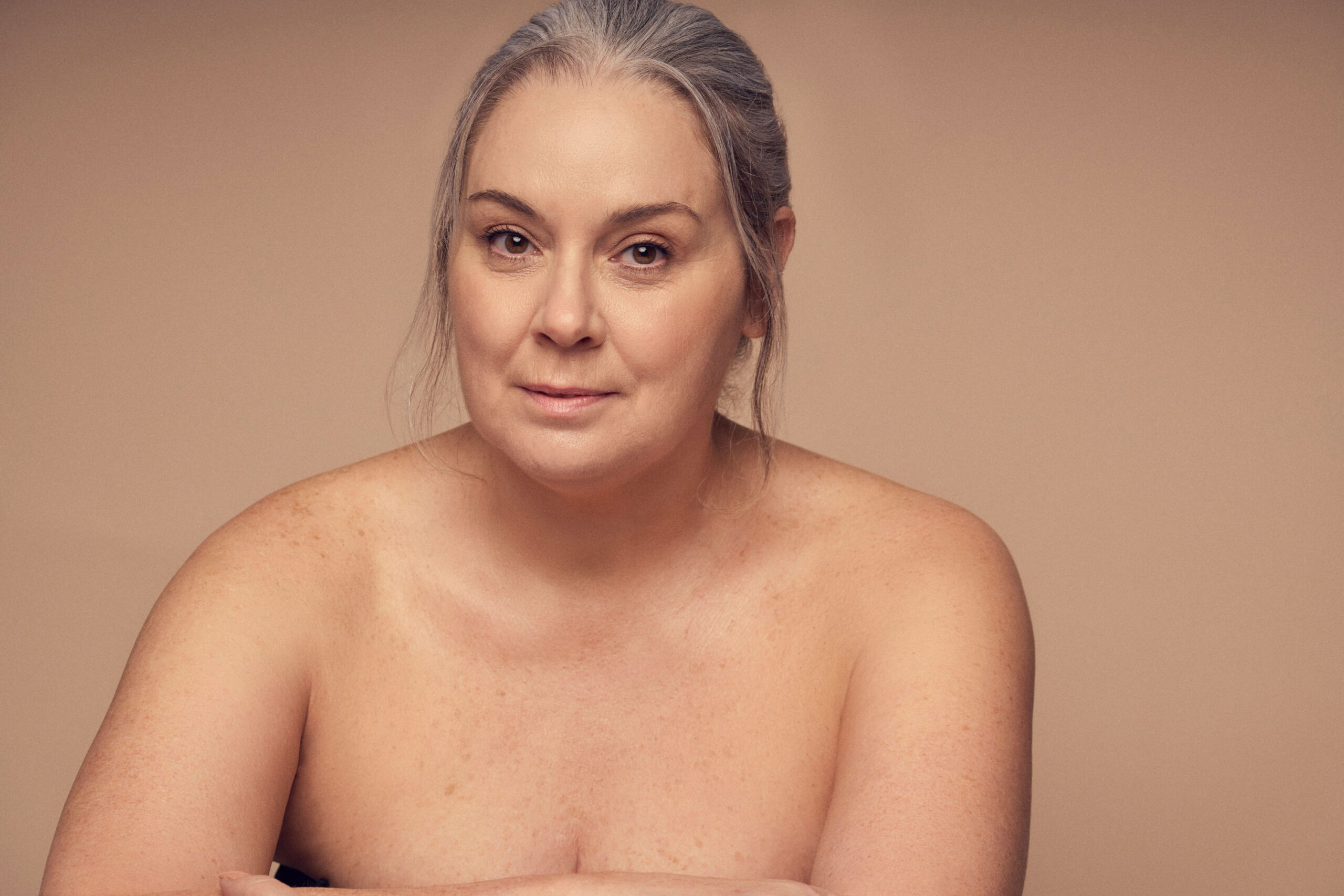 A woman with gray hair and a neutral expression sits against a plain beige background, with her arms crossed in front of her. Her skin is bare, emphasizing natural features.