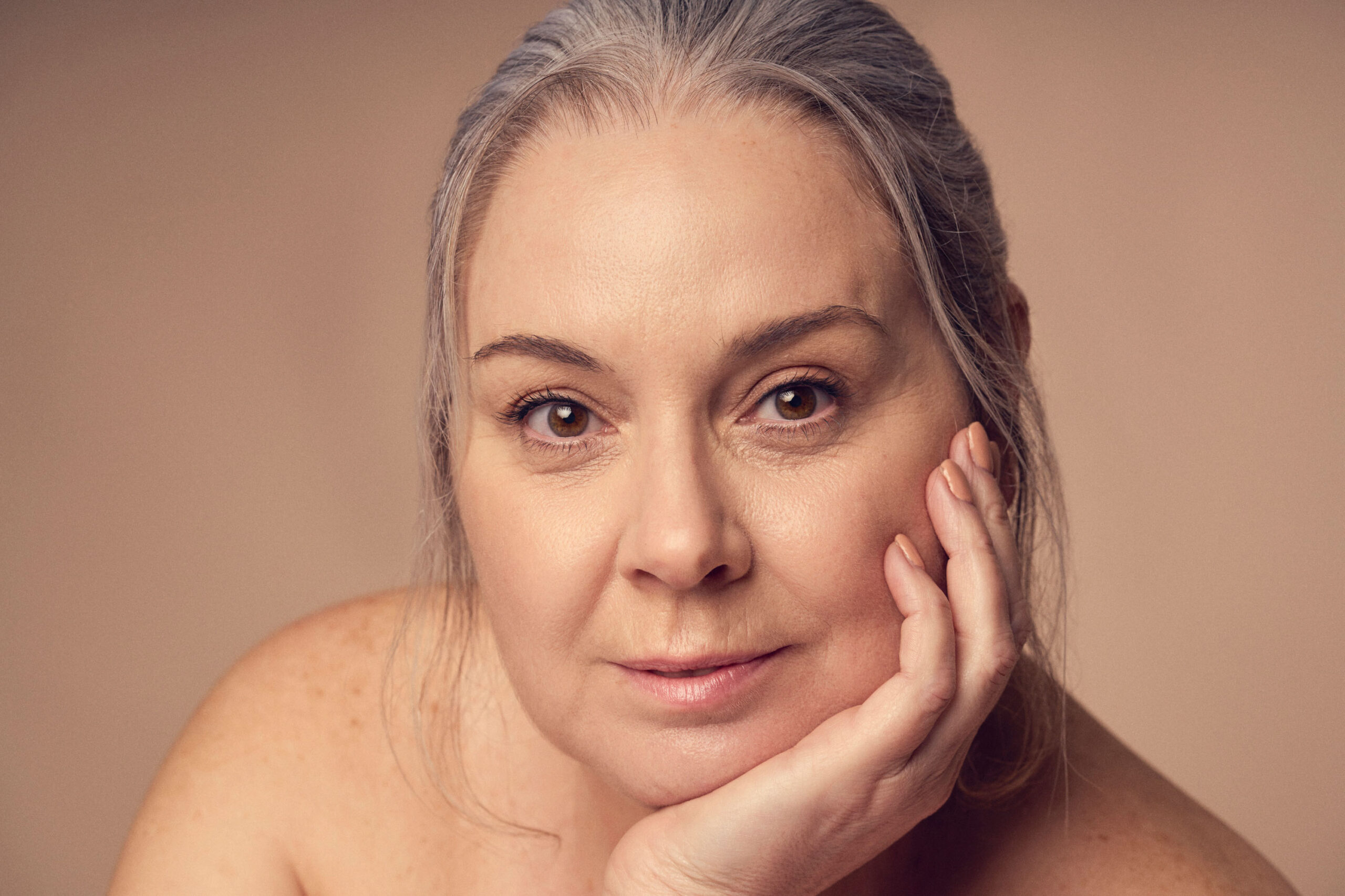 A woman with light skin and gray hair pulled back gazes softly at the camera, resting her chin on her hand. The background is a muted beige, complementing the serene and neutral expression on her face.