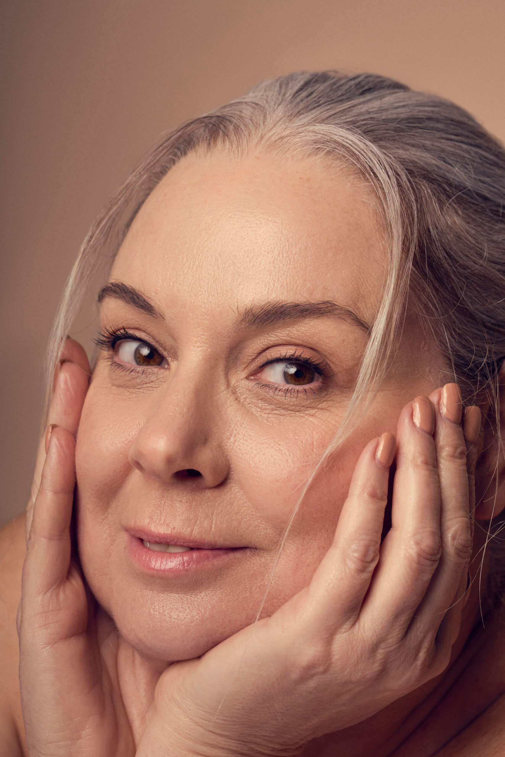 A close-up of a middle-aged person with gray hair, resting their chin on their hands. They are smiling softly, with a neutral background highlighting their relaxed expression and natural beauty.