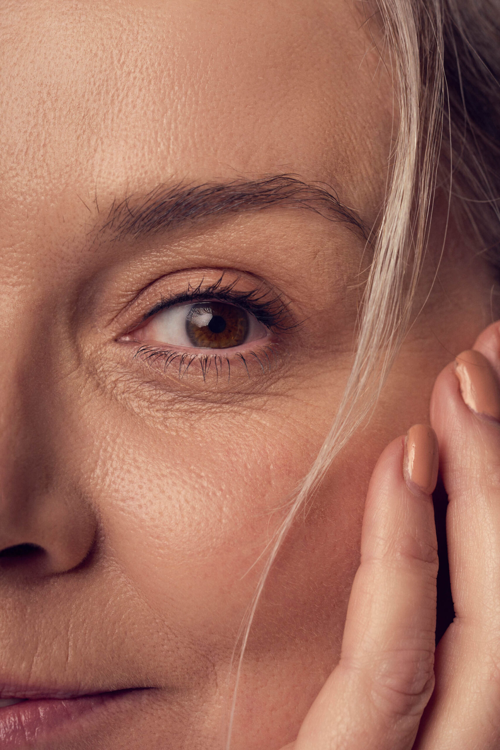 Close-up of a persons face, focusing on the eye and part of the cheek. The persons fingers gently touch the side of the face. The skin shows natural texture and some fine lines, with light makeup and neutral nail polish on the fingernails.