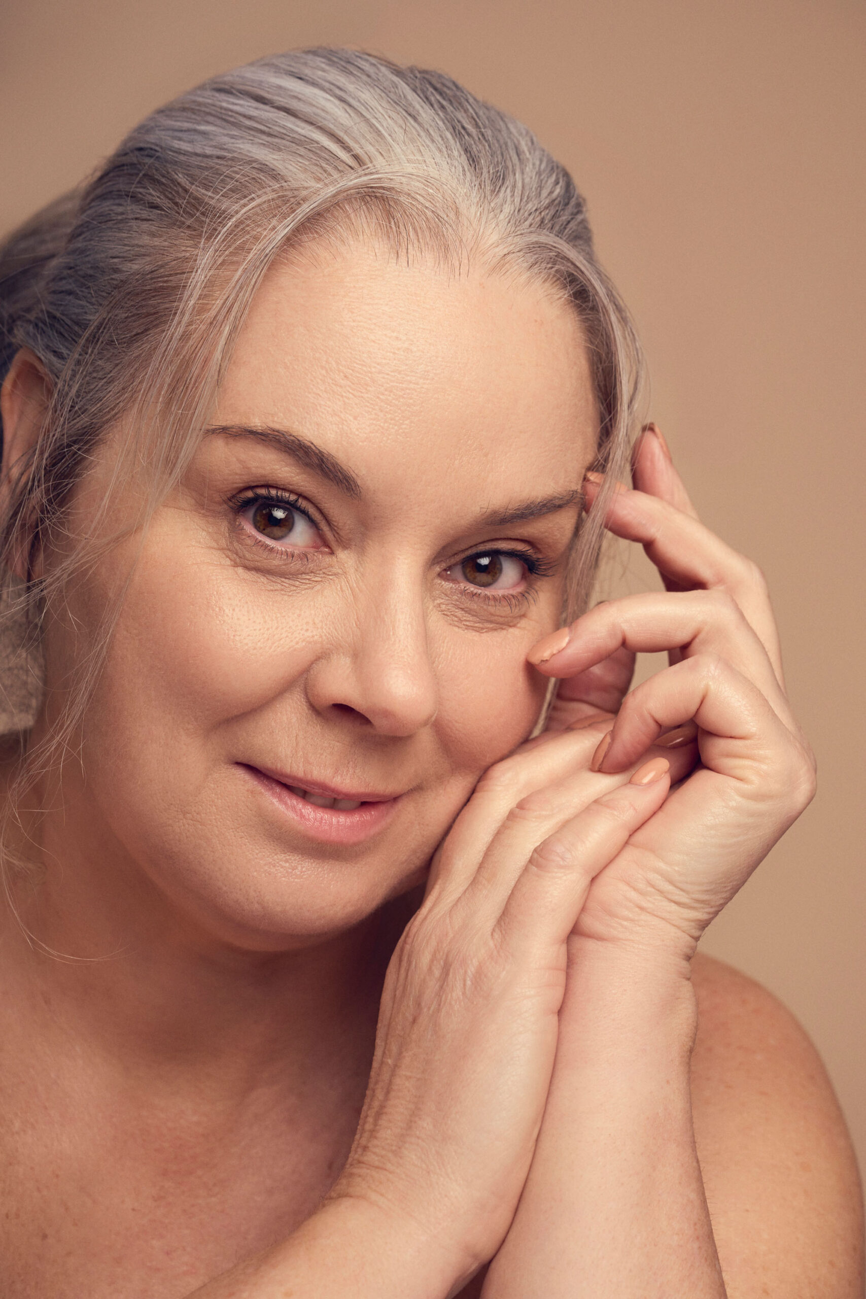 A woman with gray hair and a gentle smile rests her head on her hands. She has light-colored eyes and is posed in front of a neutral beige background. Her expression is calm and serene.