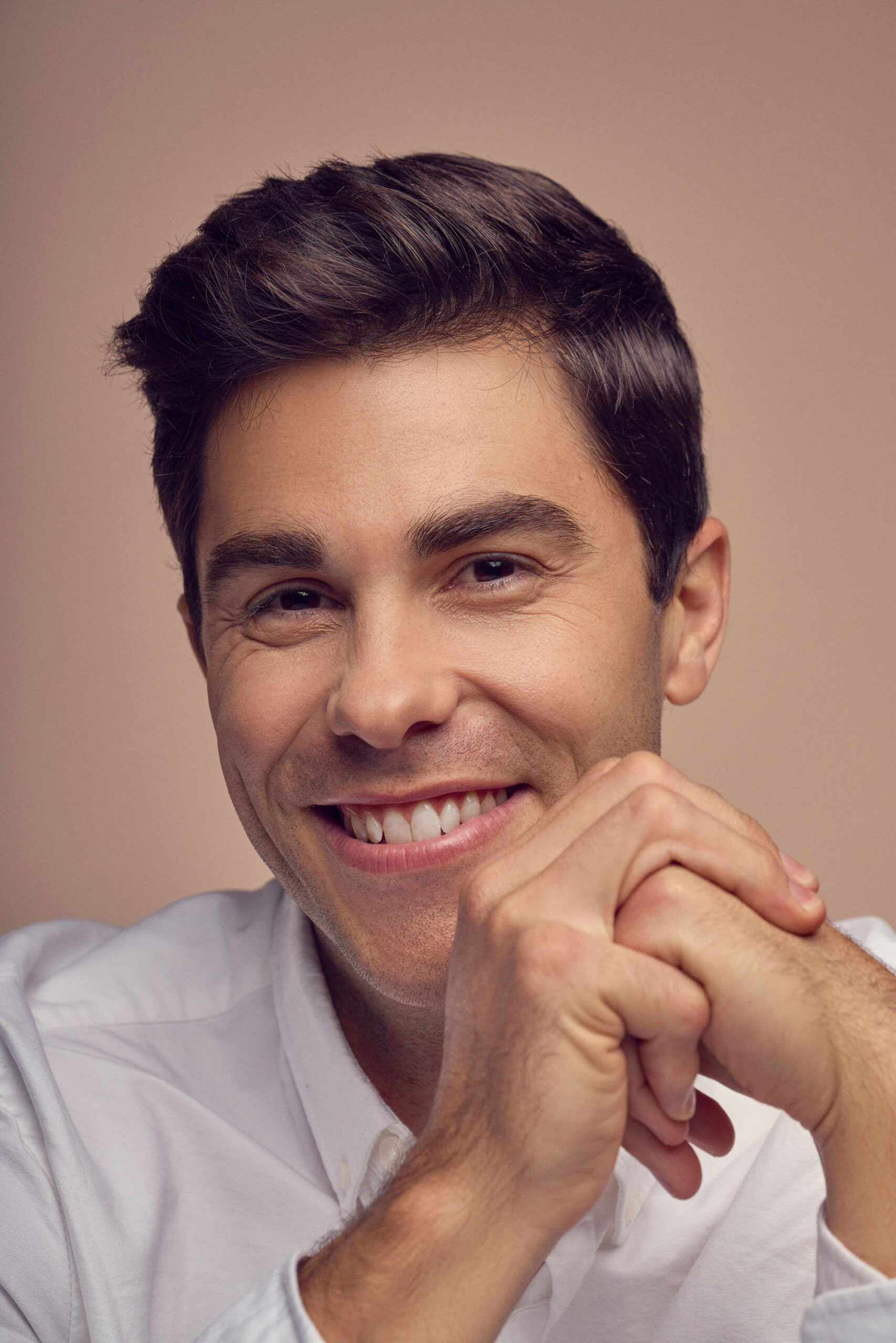 A person with short dark hair smiling softly, wearing a white shirt, rests their chin on their clasped hands against a neutral background.