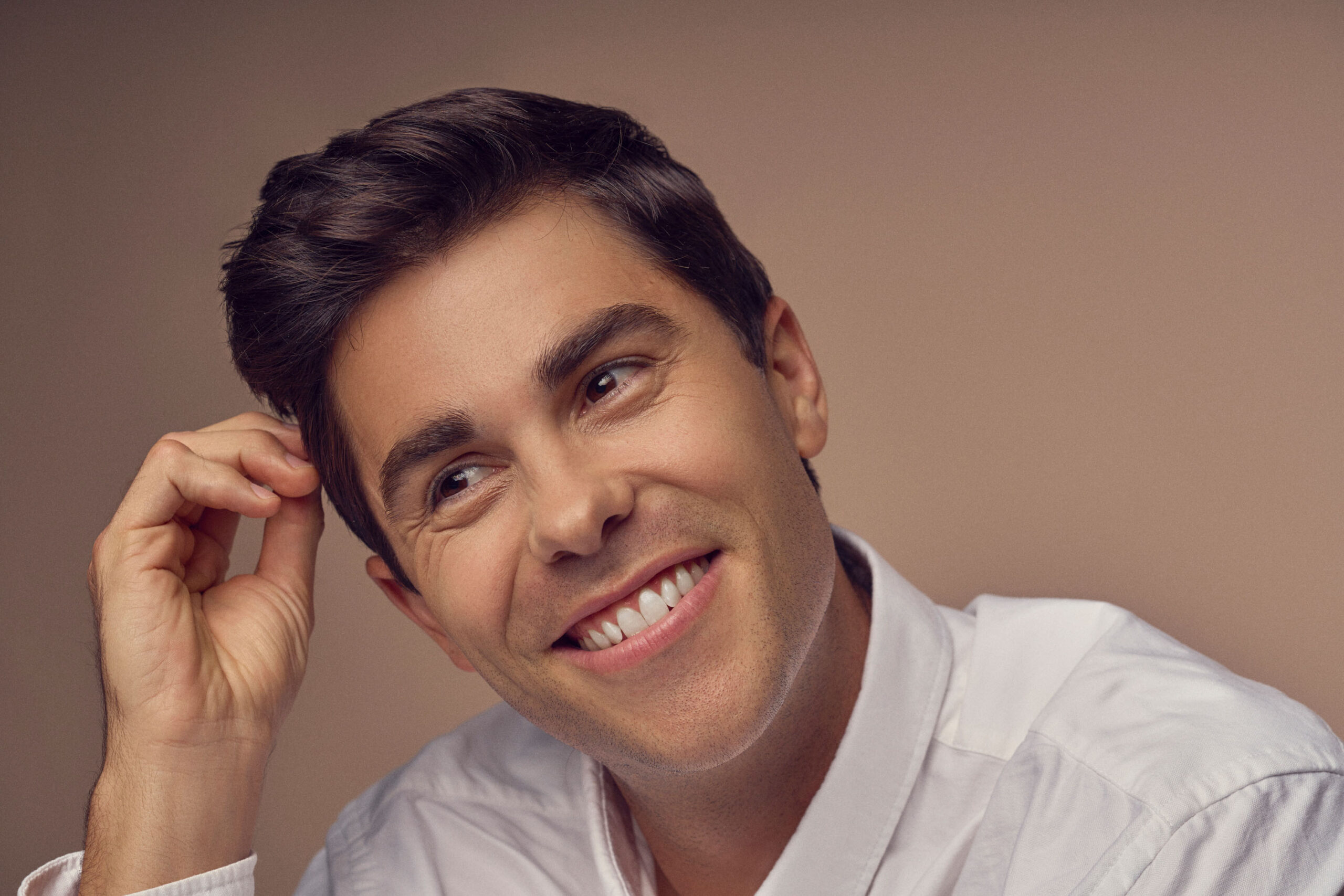 A person with dark hair and a friendly smile is seen wearing a white shirt. They are touching their hair while looking slightly upward against a neutral background.