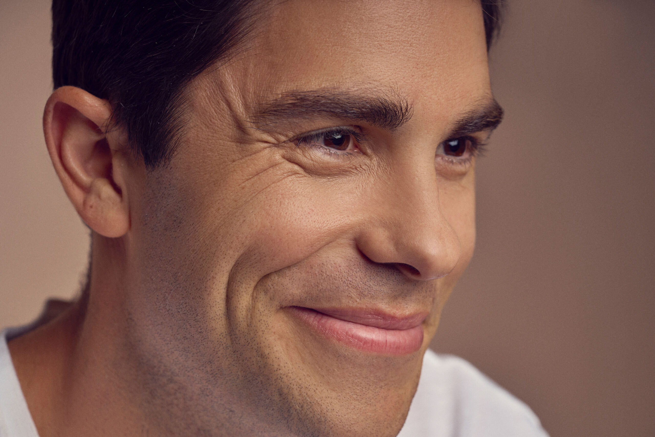 A person with short dark hair smiling warmly, wearing a light-colored shirt. The background is blurry, emphasizing the subjects facial features and expression.