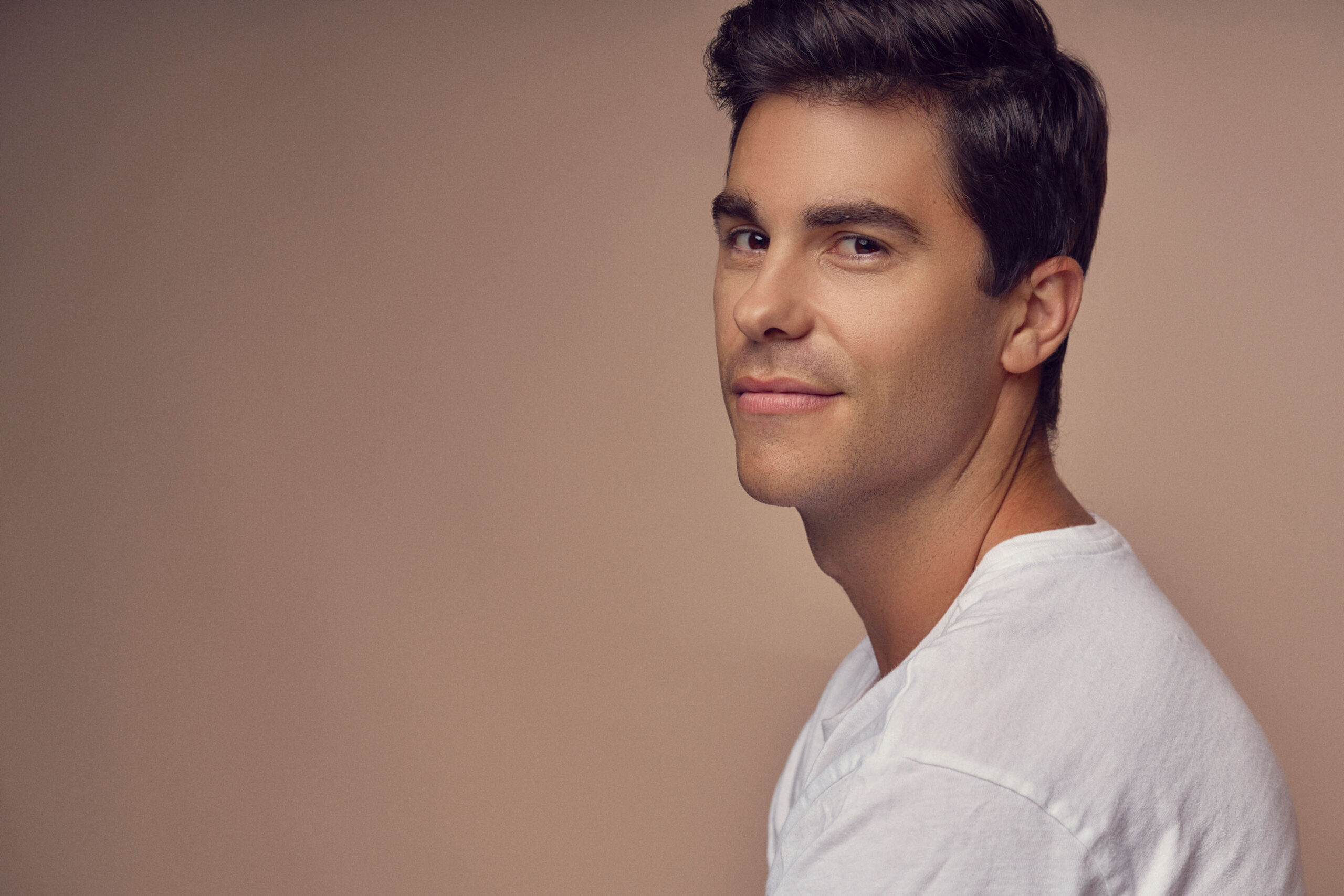 A man with short dark hair is wearing a white T-shirt. He is smiling slightly and looking towards the camera against a neutral beige background.