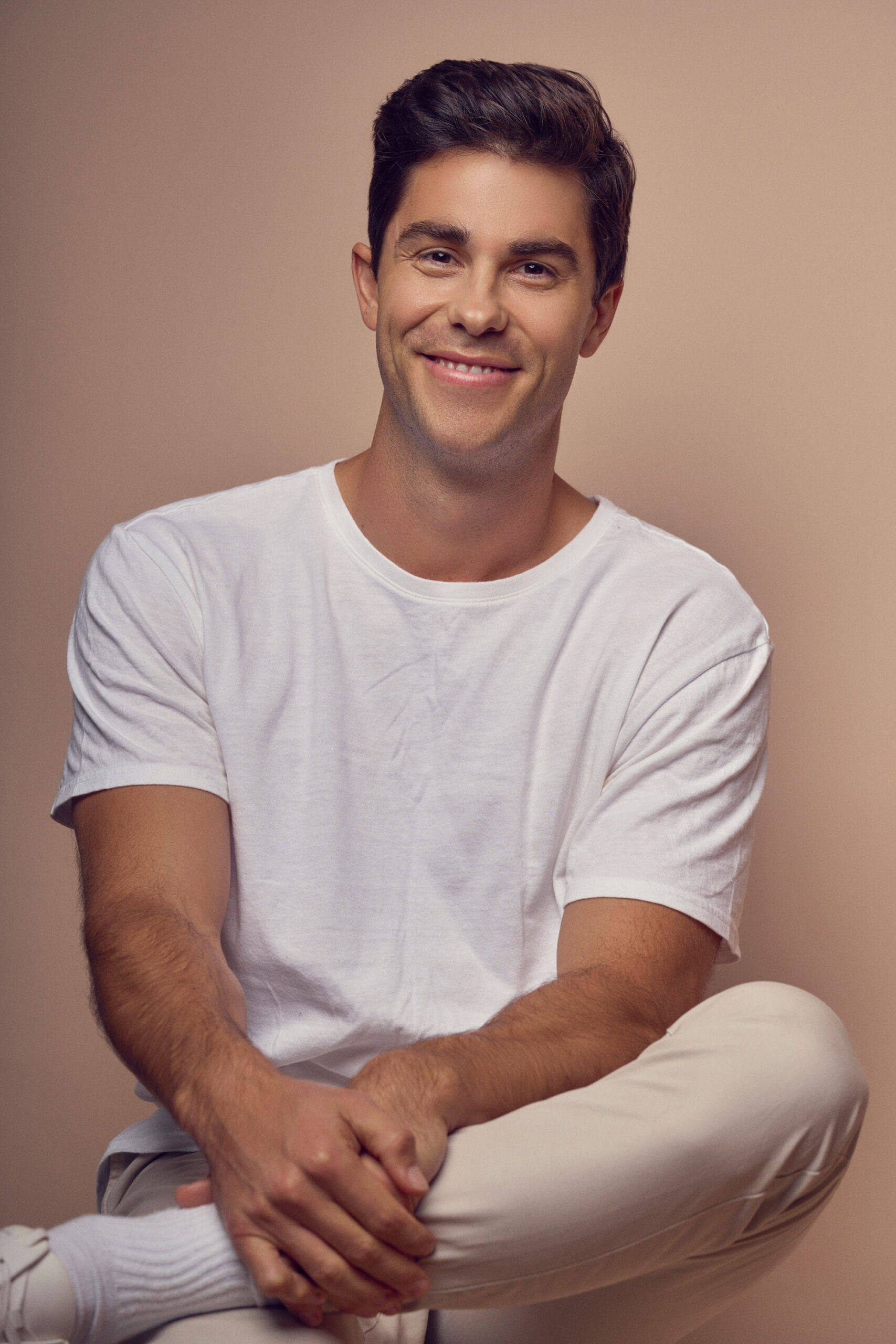 A man with short dark hair sits with a relaxed pose, wearing a white t-shirt and light-colored pants. He is smiling and seated against a plain, neutral background.