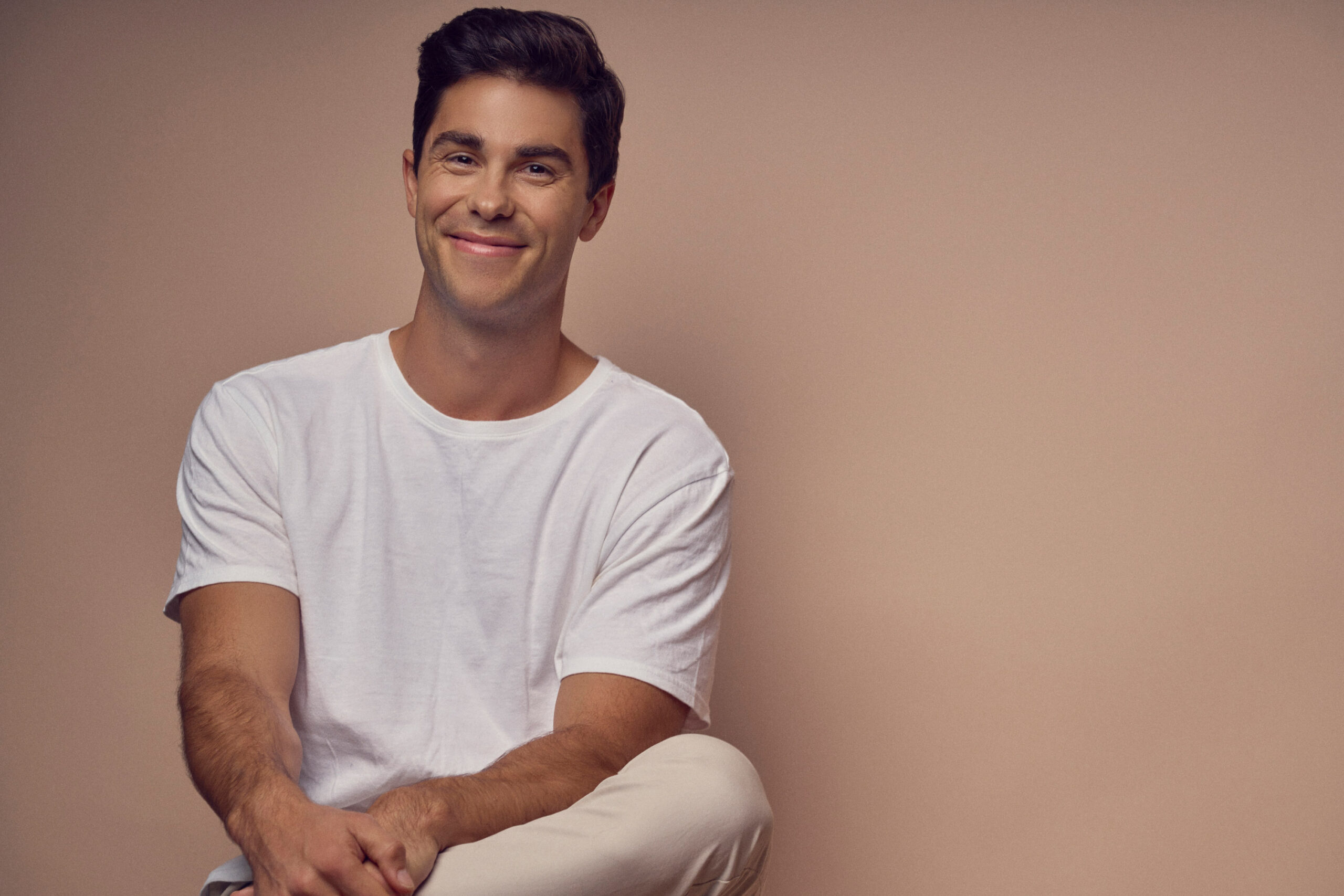 Smiling man in a white t-shirt sits against a neutral beige background.