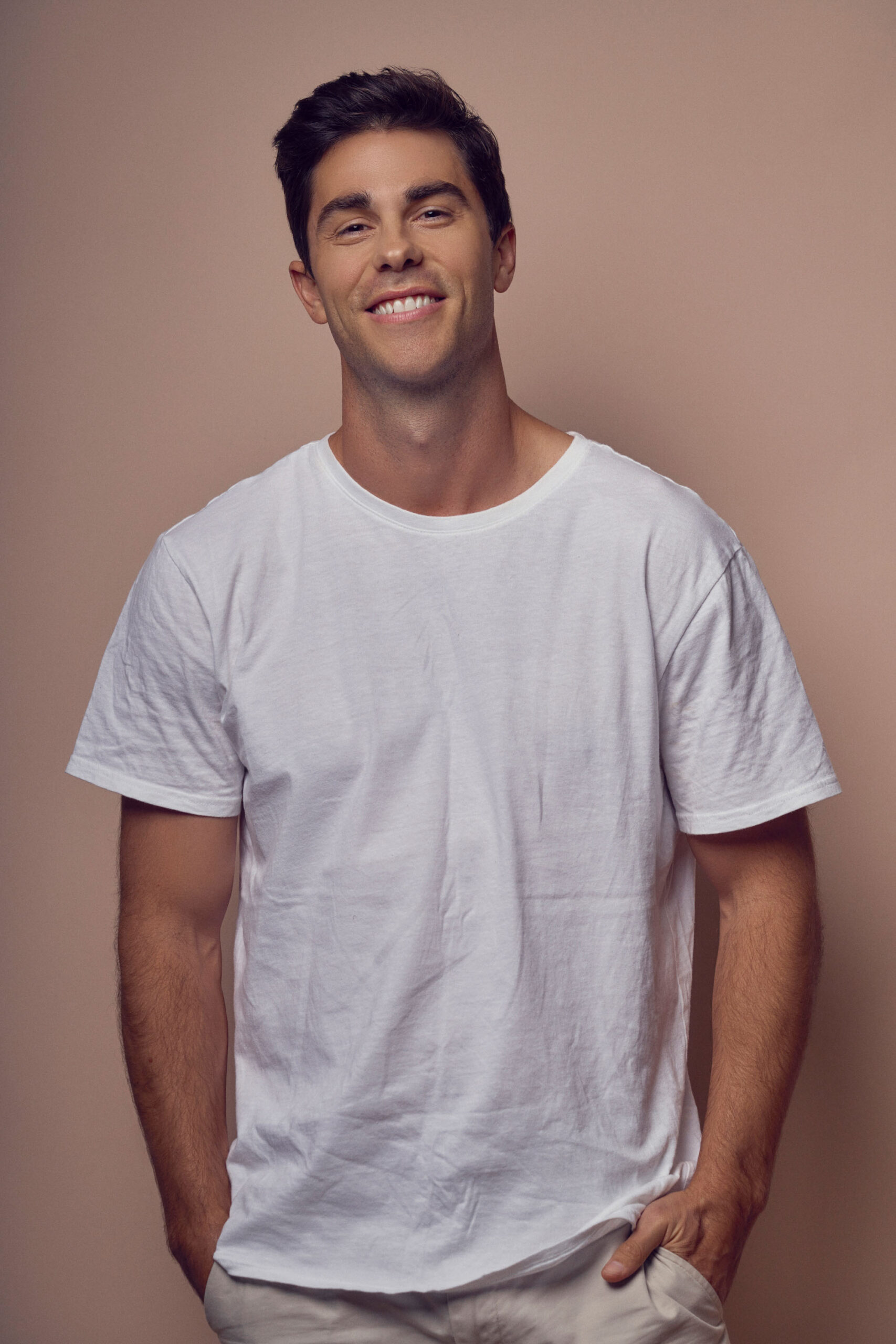 A man wearing a white t-shirt stands against a plain beige background, smiling at the camera with his hands in his pockets. He has short dark hair and appears relaxed and cheerful.