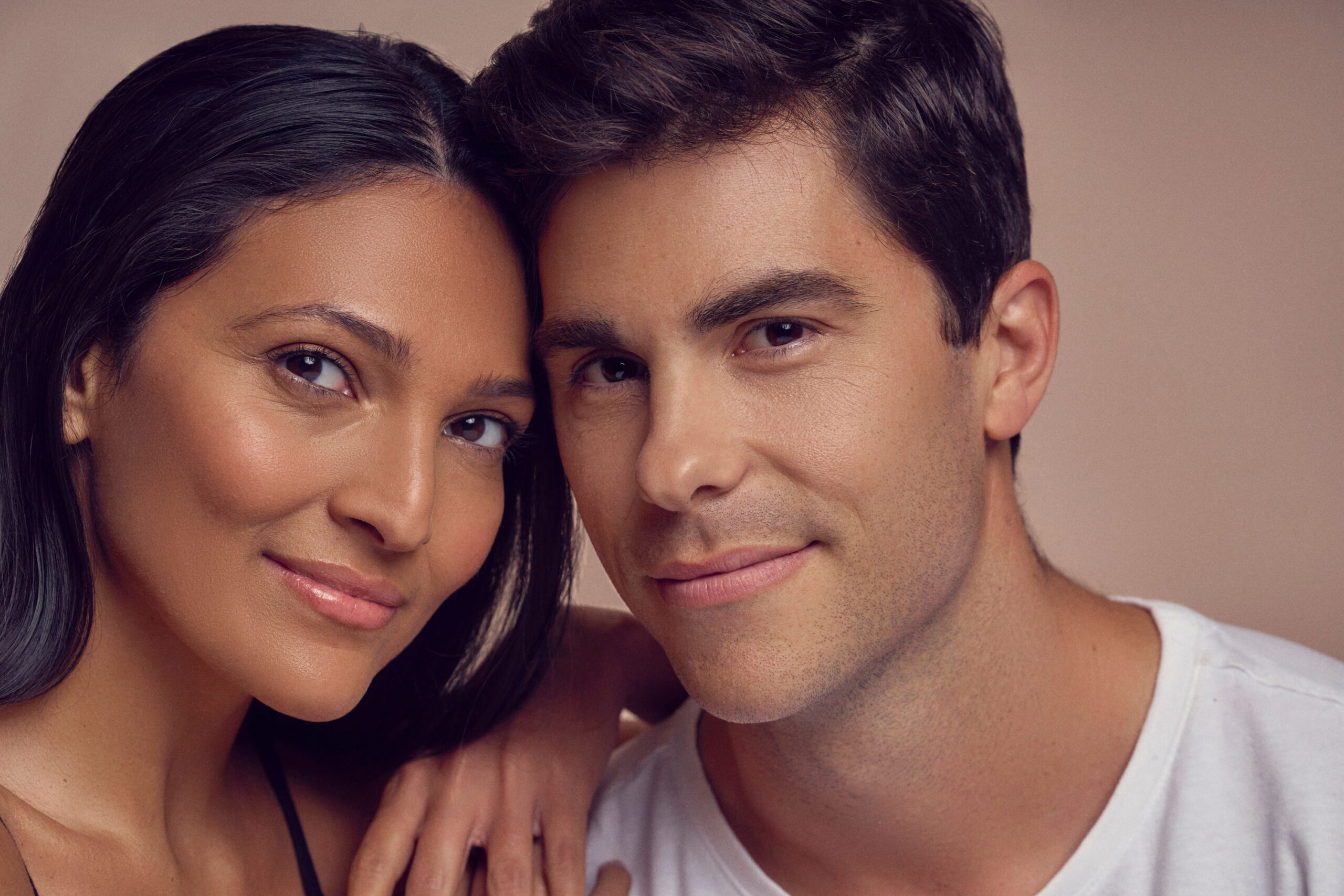 A close-up image of a smiling couple. The woman has long dark hair and the man has short dark hair. They both wear casual tops and share a warm expression against a neutral background.