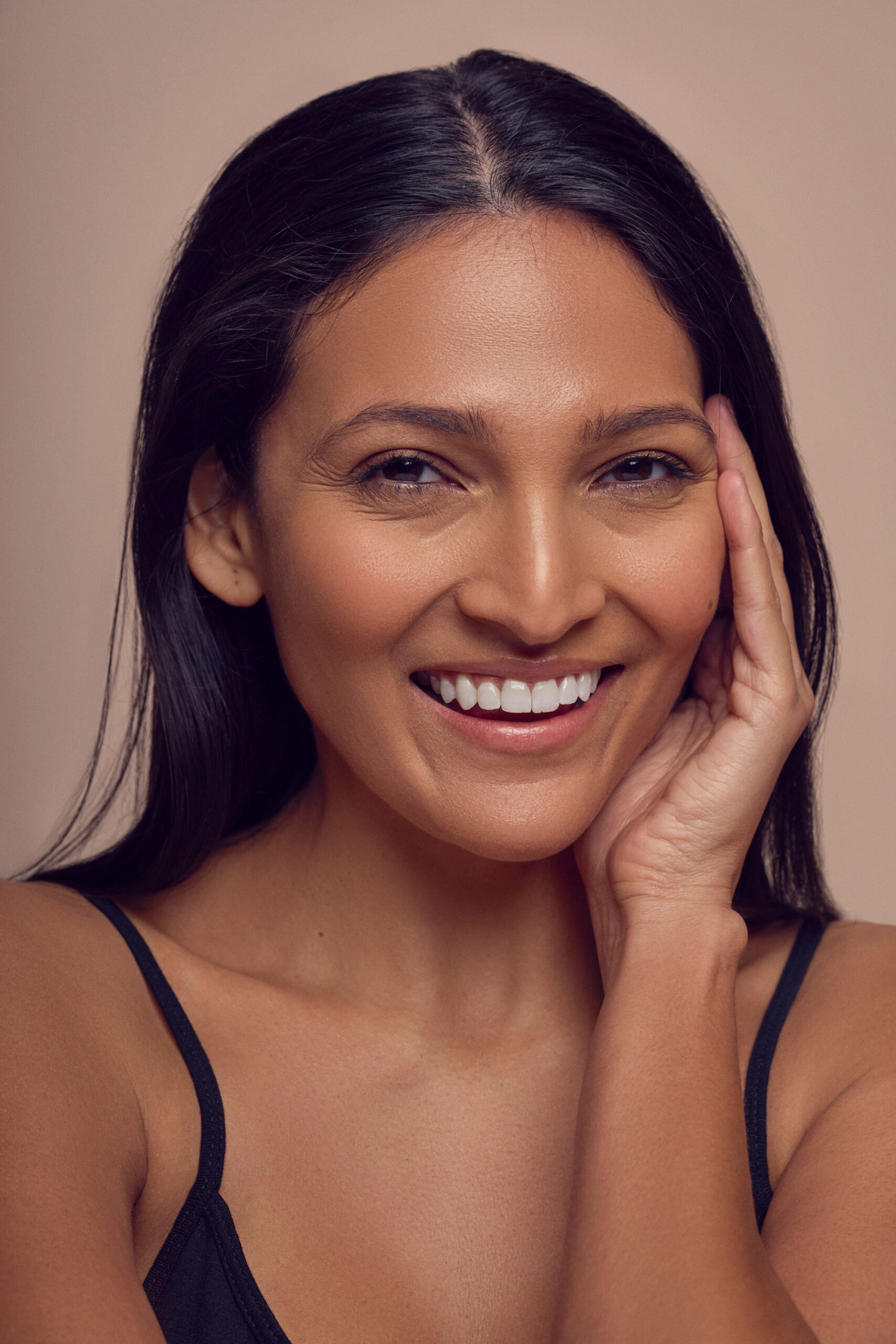 A person with long dark hair smiles warmly, holding their right hand to their face. They wear a black top against a neutral background, creating a soft, friendly portrait.