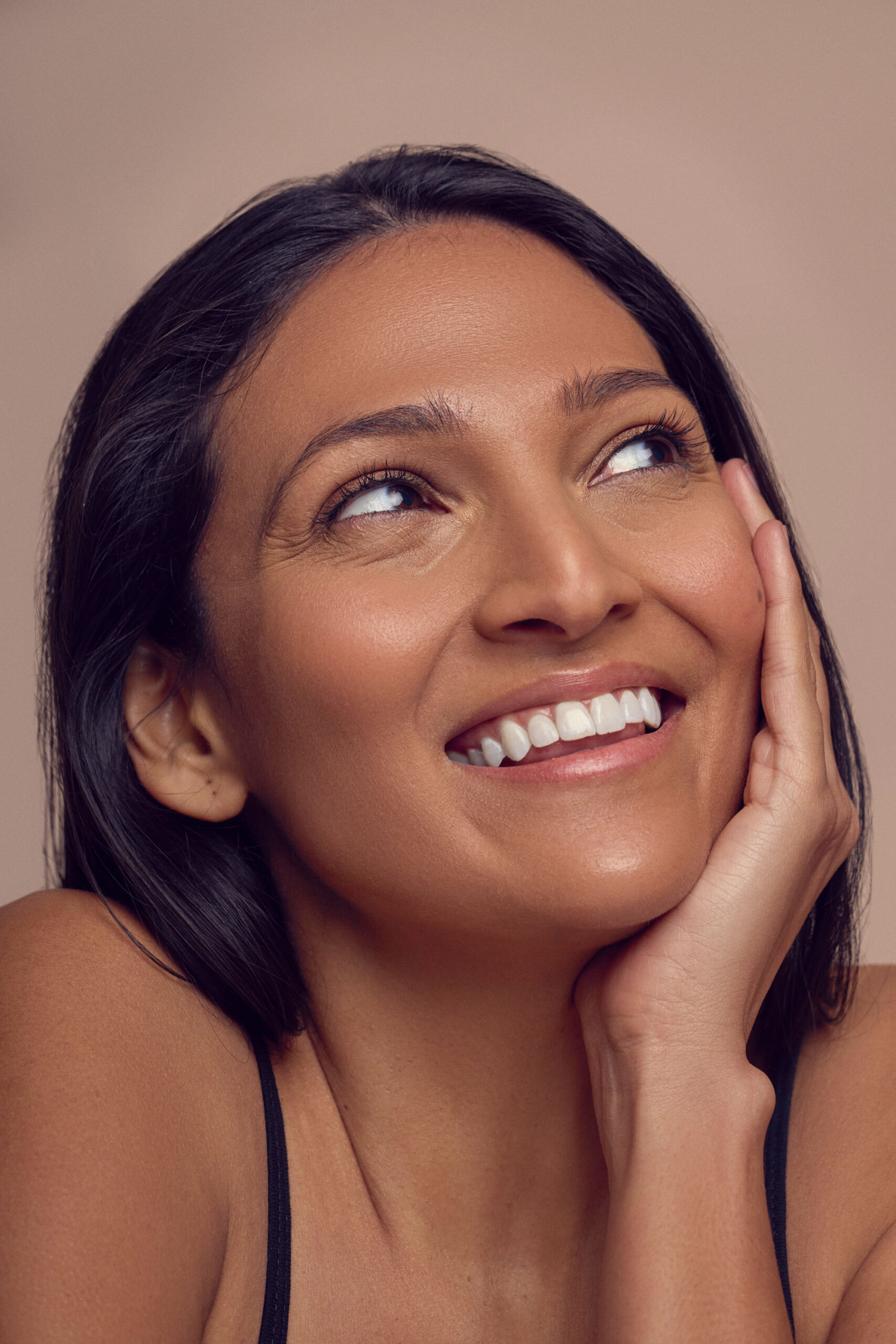 A woman with long dark hair smiles gently, looking upwards with her head resting on her hand. She has a light complexion and wears a black tank top. The background is a soft beige color.