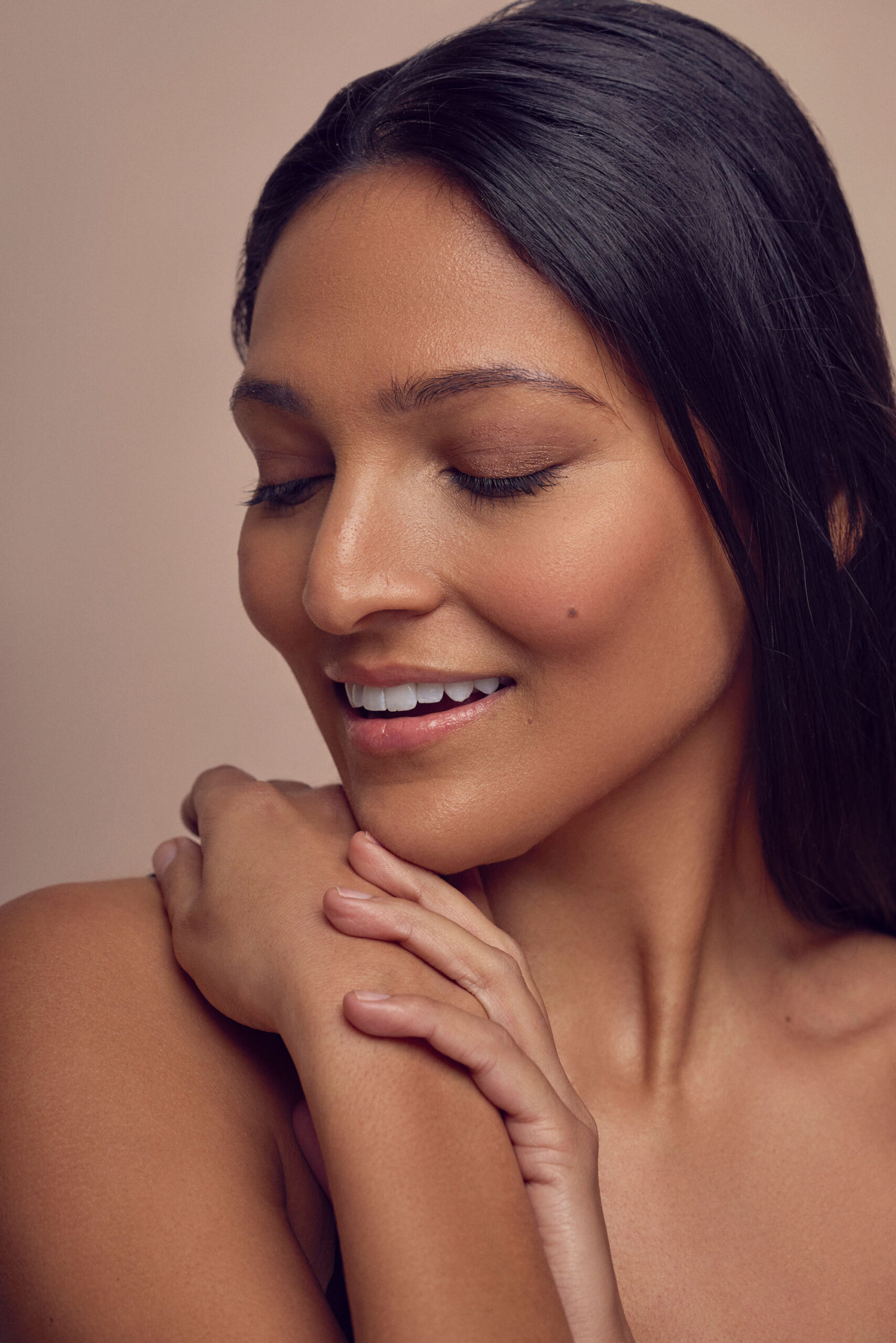 A person with long dark hair smiles gently with eyes closed. They rest a hand on their shoulder, displaying manicured nails. The background is a soft, neutral tone, creating a serene atmosphere.