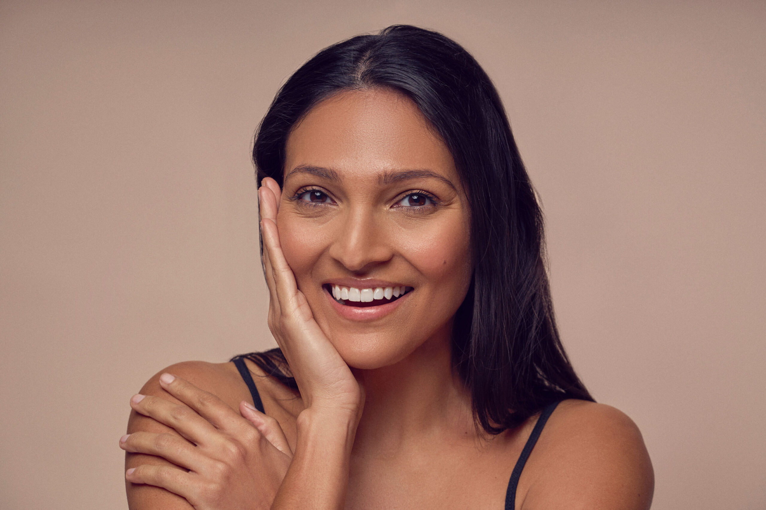 A woman with long dark hair smiles warmly, resting her hand on her cheek. She is wearing a black tank top and has a neutral background. The image conveys a sense of happiness and confidence.