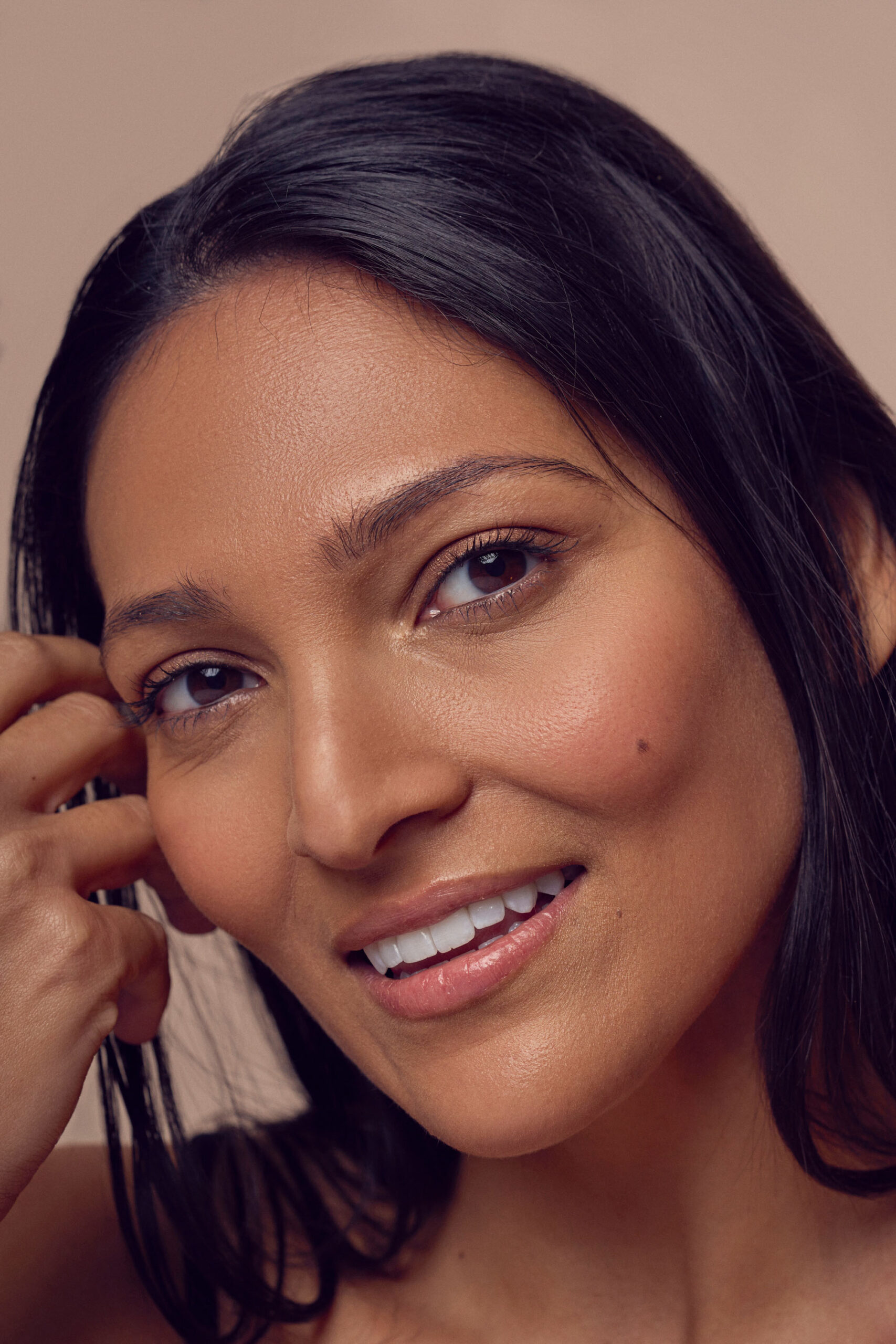 A woman with long dark hair smiles gently. She has clear skin and is touching her hair with one hand. The background is a soft beige, complementing her natural complexion.