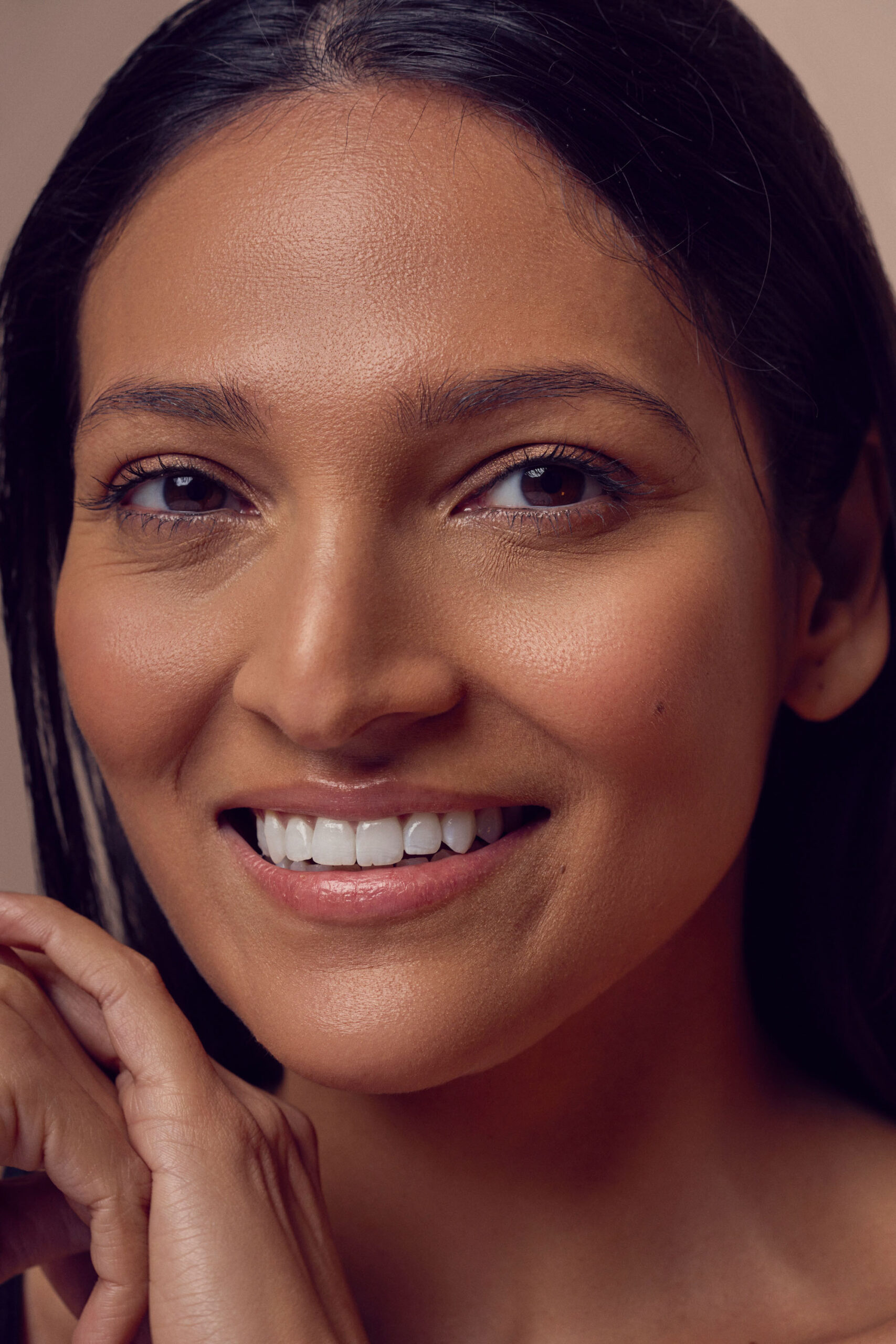 A woman with long, dark hair smiles warmly at the camera. Her hands are gently positioned near her chin. The background is a soft, neutral color, highlighting her natural, radiant complexion.