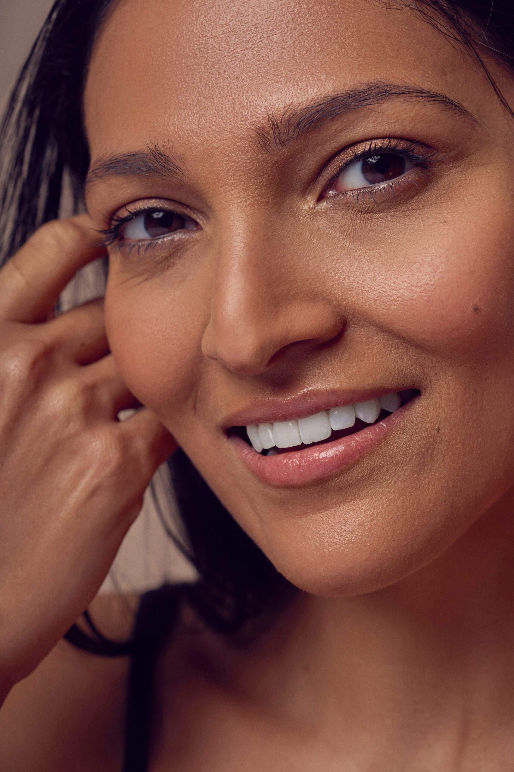 A close-up of a person with long dark hair, smiling, and gently touching their face with their fingers. Their skin appears smooth, and they have a natural, relaxed expression.