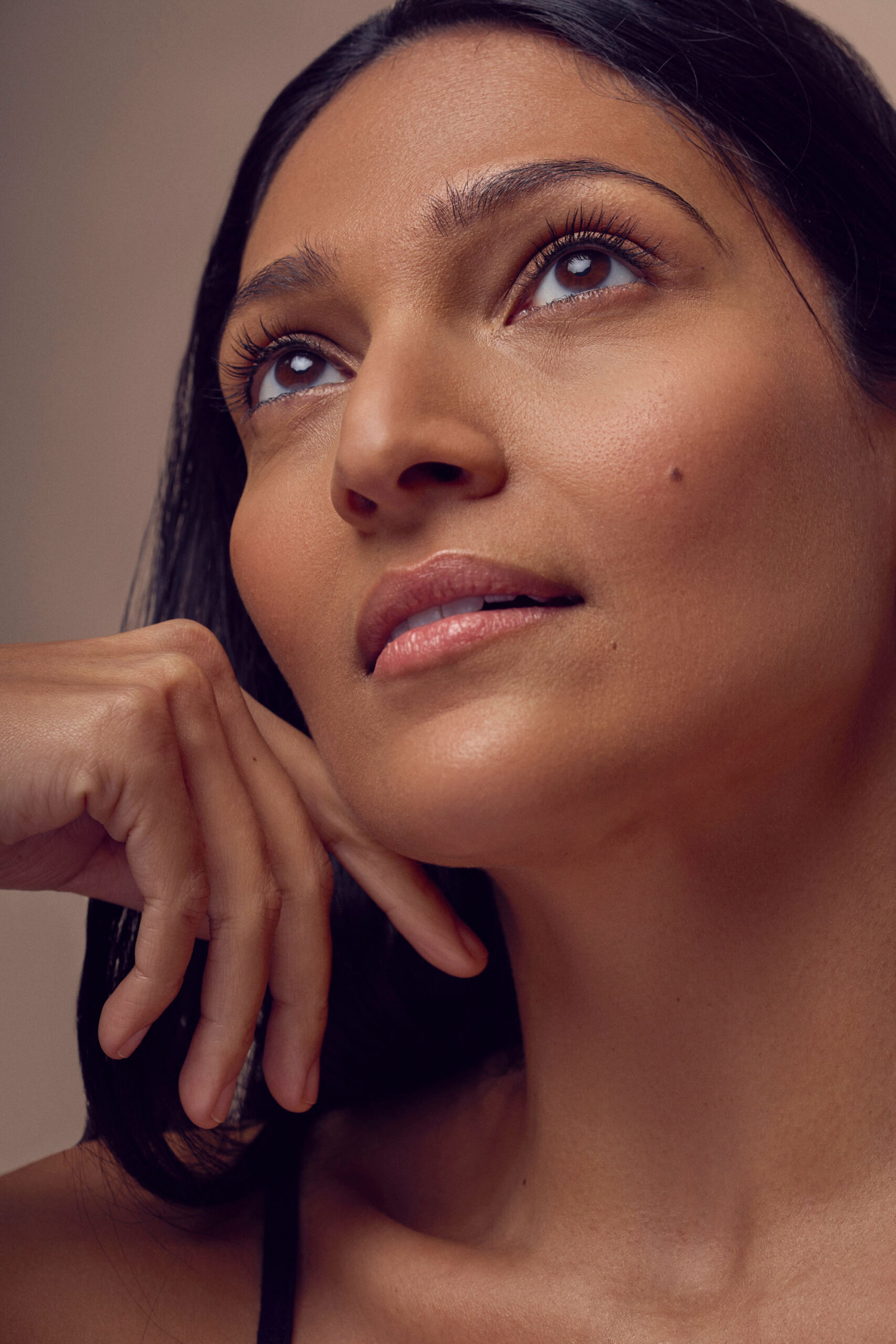 A person with long dark hair gazes upward with a thoughtful expression. They rest their chin on their hand, showcasing smooth, even-toned skin and natural makeup. The lighting is soft, highlighting their facial features.