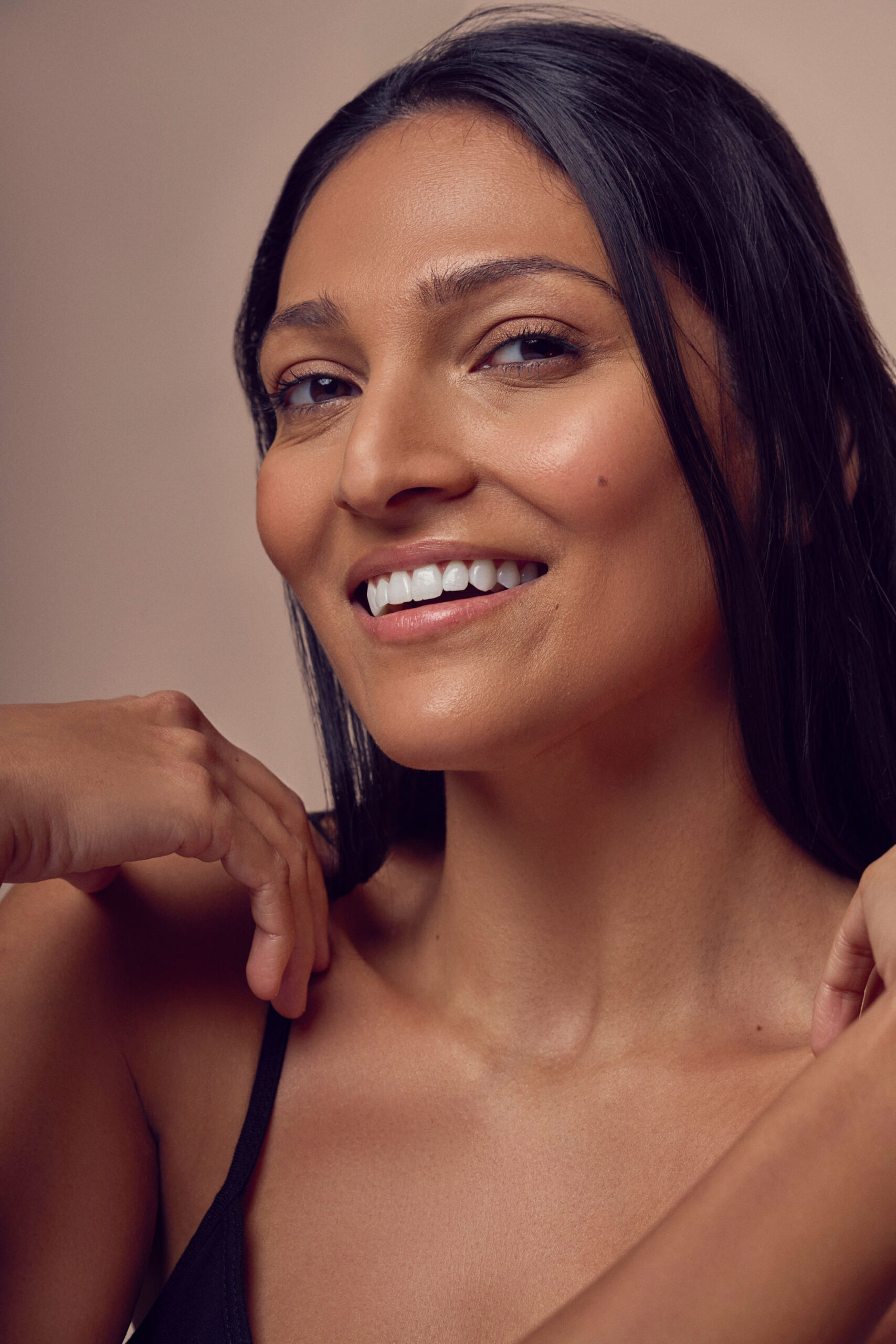 A woman with long dark hair smiles warmly. She has clear skin and bright white teeth, wearing a black sleeveless top. Her hands are gently placed near her face against a soft, neutral background.