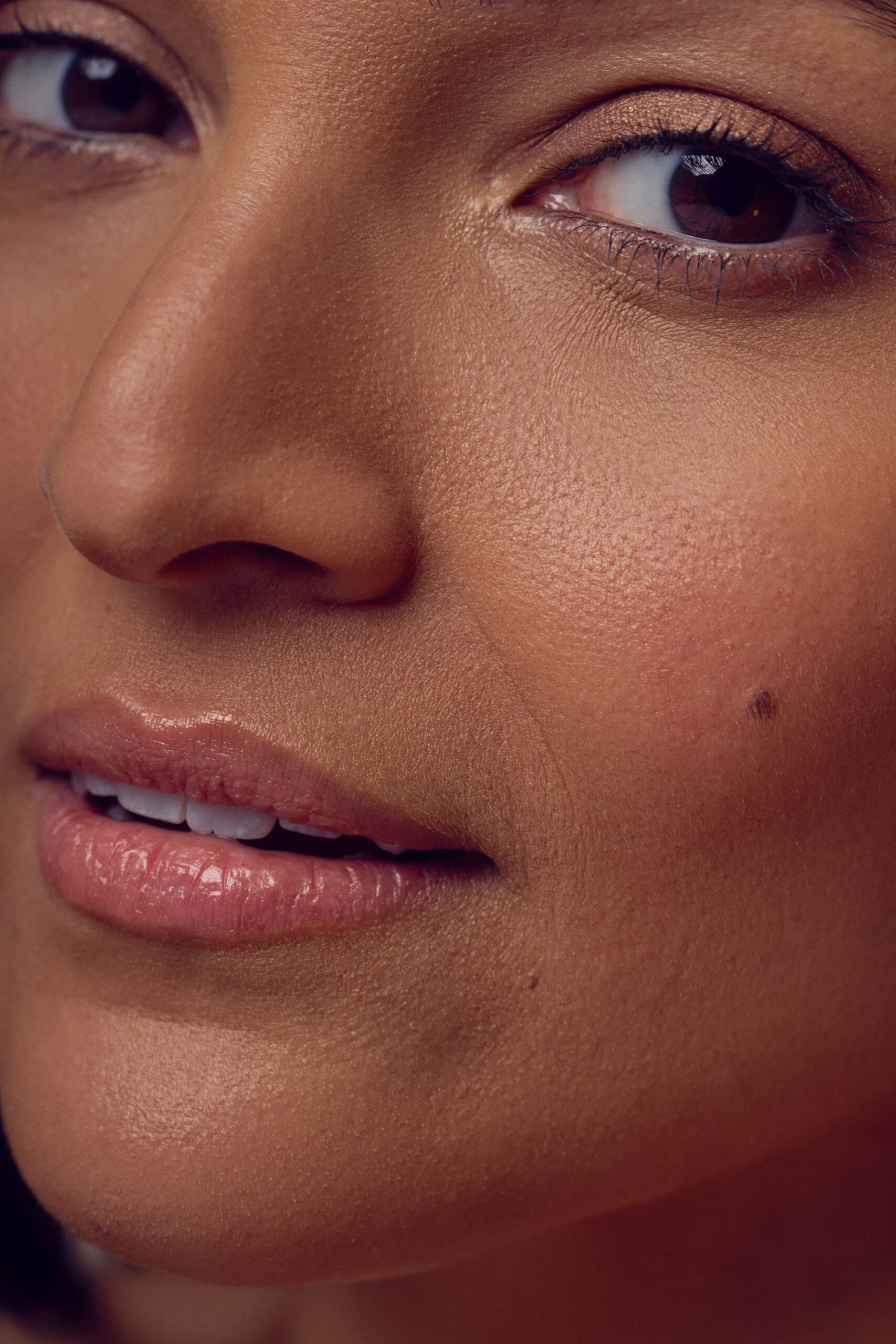 Close-up of a womans face focusing on her eyes, nose, and mouth. Her skin has a natural, smooth texture, with a small beauty mark on her cheek. Her lips are slightly parted, and her eyes are softly gazing at the camera.