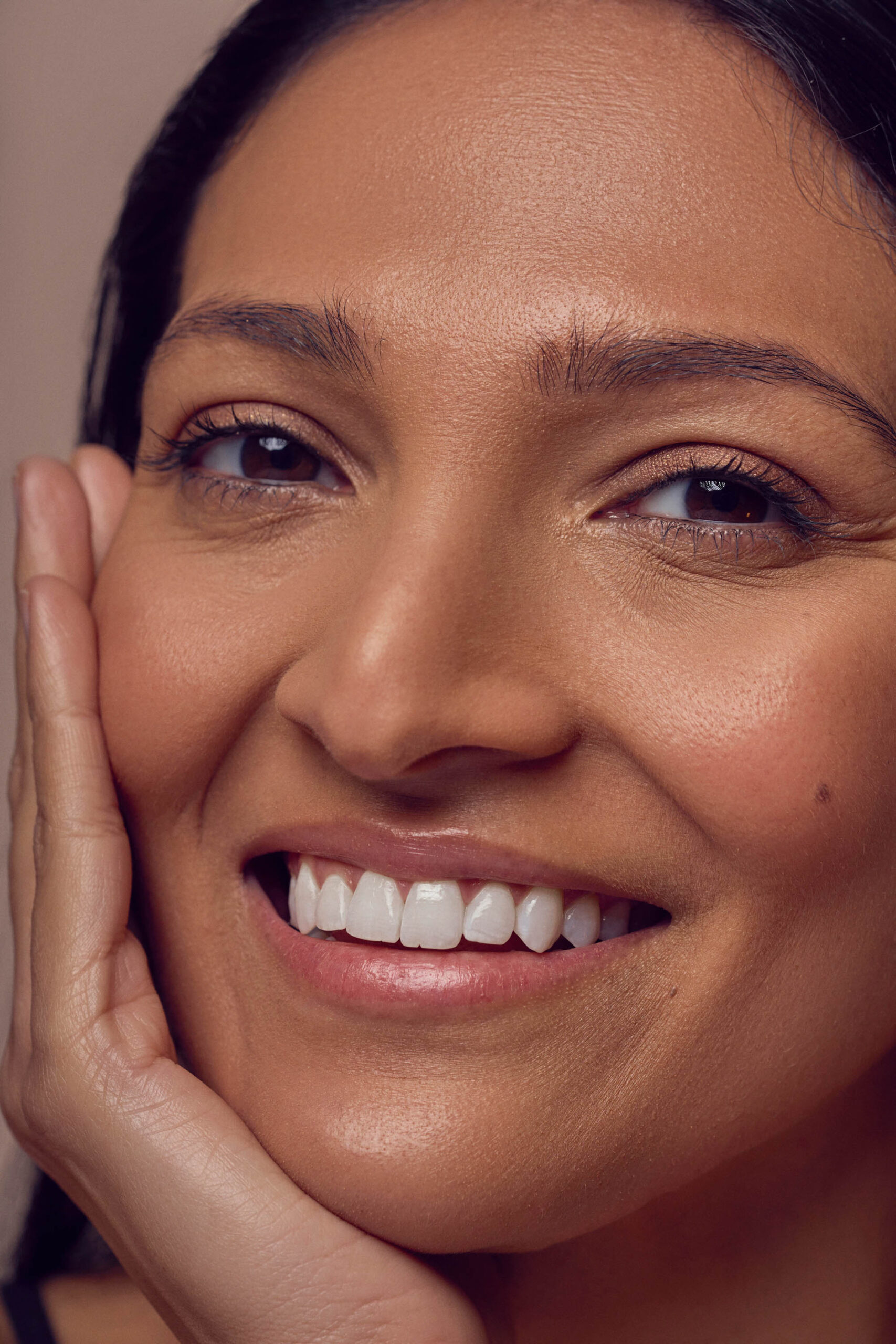 A close-up of a person smiling, with long dark hair and radiant skin. Their hand rests gently on their cheek, highlighting their bright eyes and white teeth.