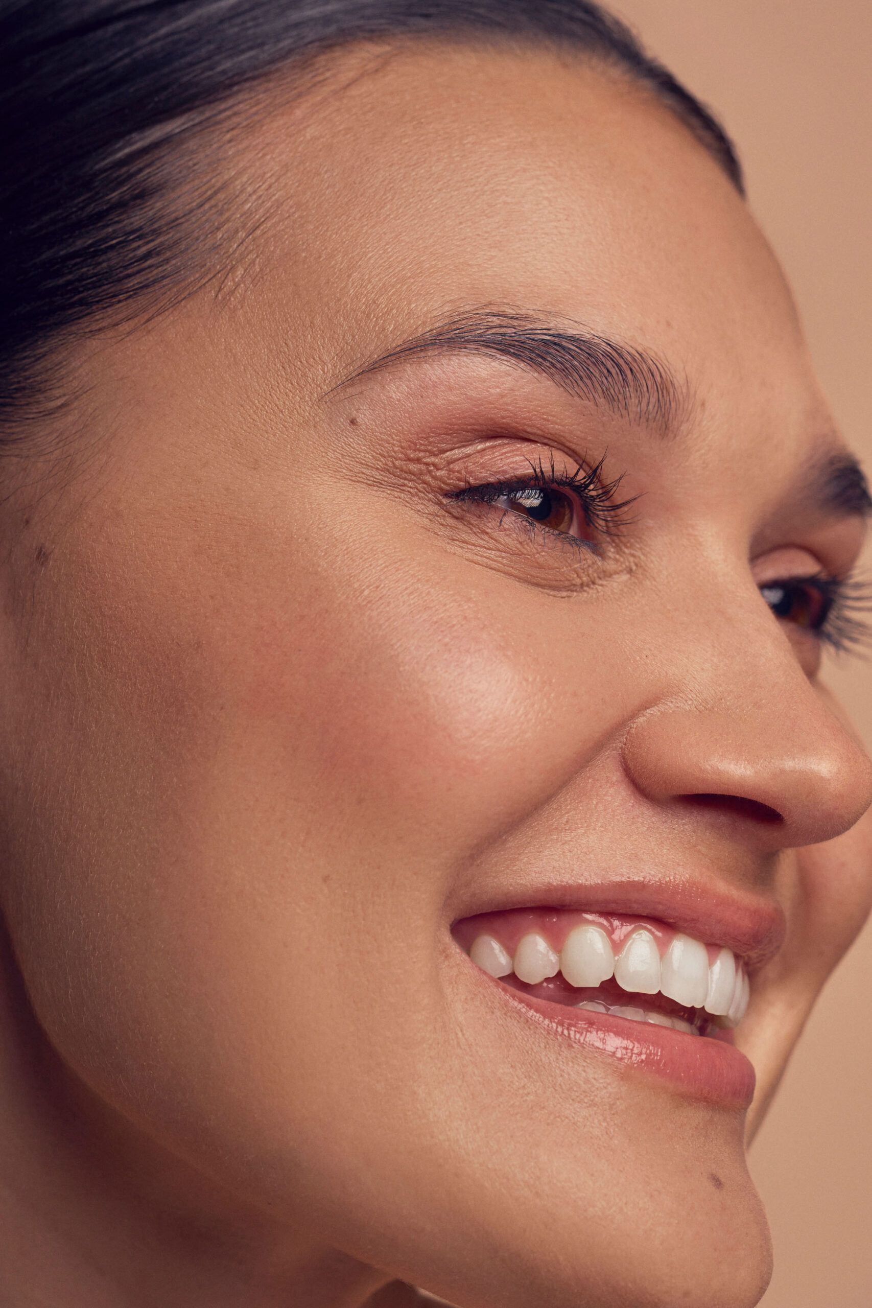 Close-up of a person smiling with a clear, glowing complexion. Their dark hair is pulled back, and they have prominent eyelashes and well-defined eyebrows. The background is a soft, neutral tone.