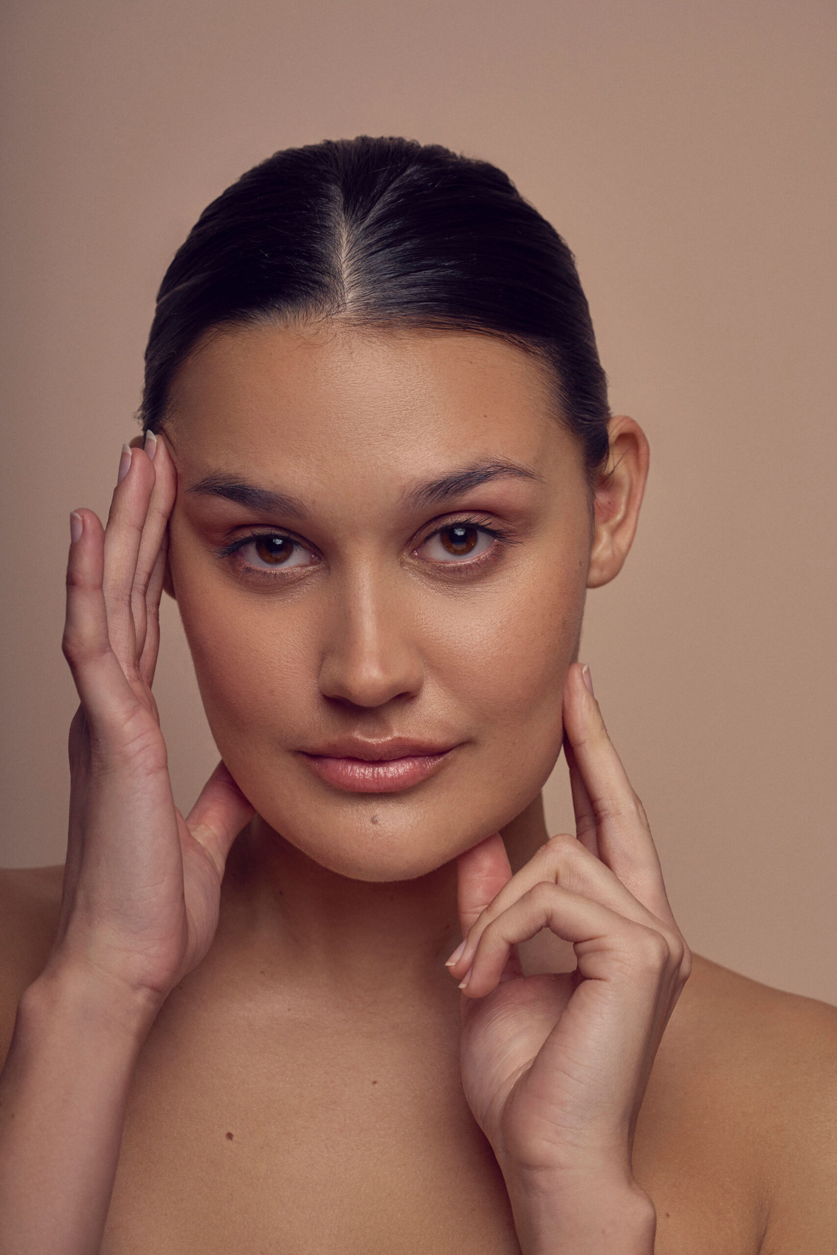 A person with slicked-back dark hair poses in front of a neutral background. They hold one hand gently on their cheek and another near their jawline, looking directly at the camera with a calm expression.