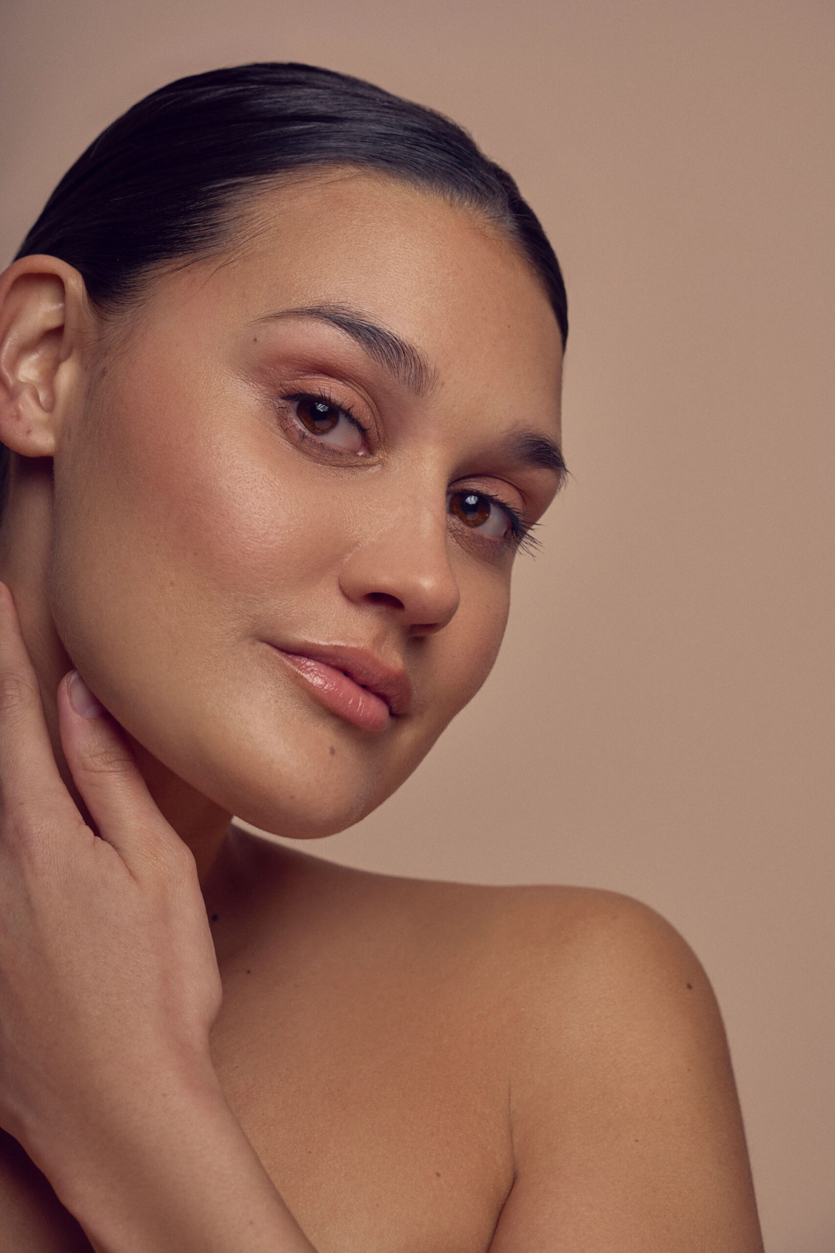 A person with smooth skin and dark hair is posing against a neutral background. They have a serene expression and are touching their neck with one hand, showcasing a natural makeup look.