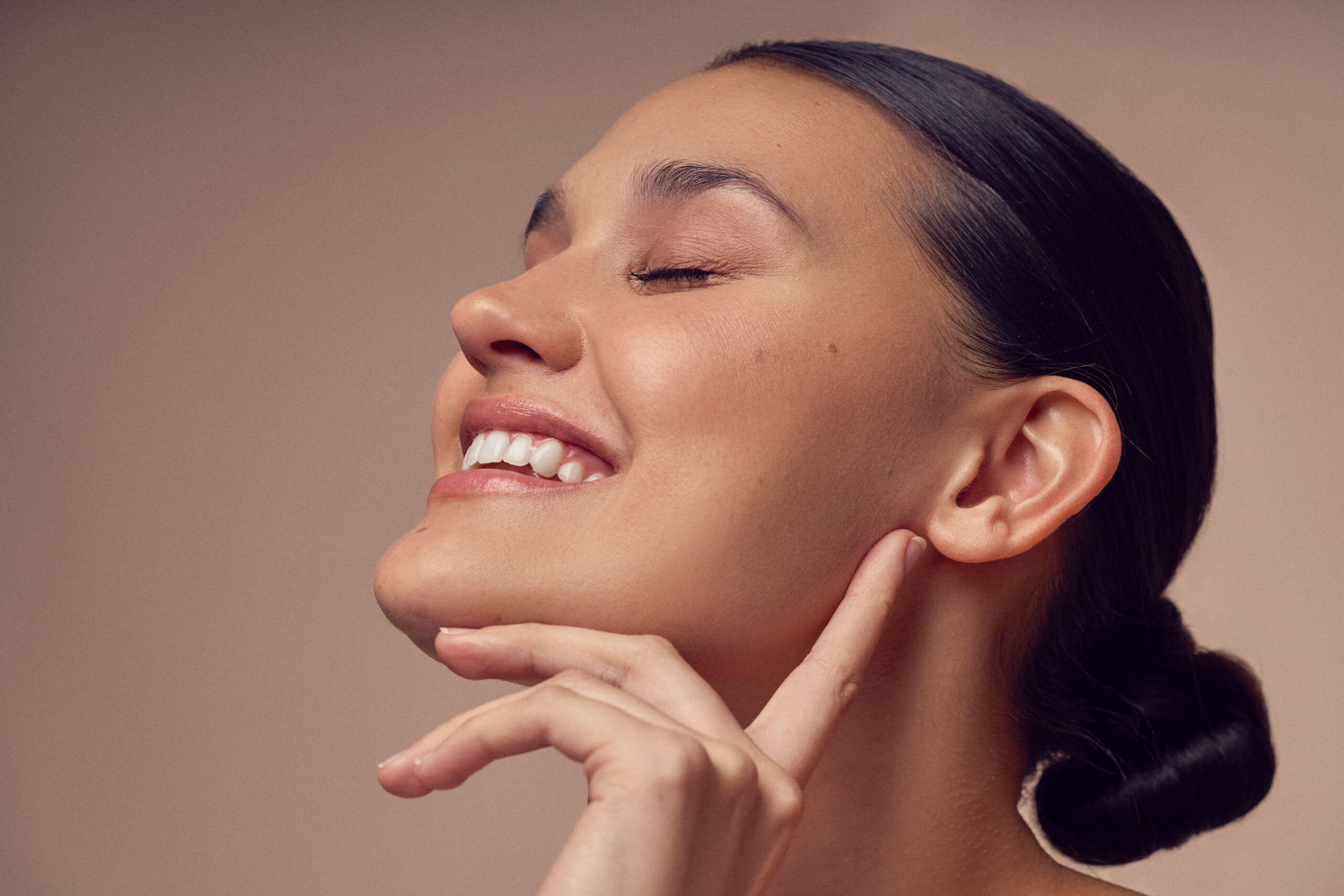 A woman with her hair tied back enjoys a serene moment with her eyes closed and a gentle smile. She touches her face lightly with a hand, set against a soft beige background.